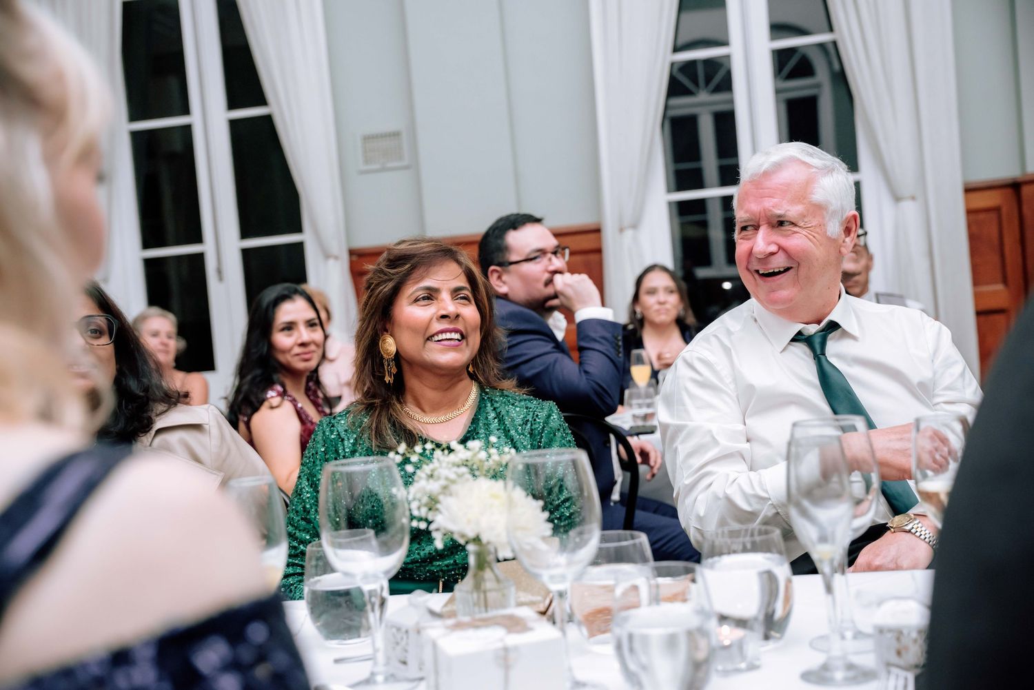 Wedding guests laughing and applauding during heartfelt speeches at elegant reception dinner.