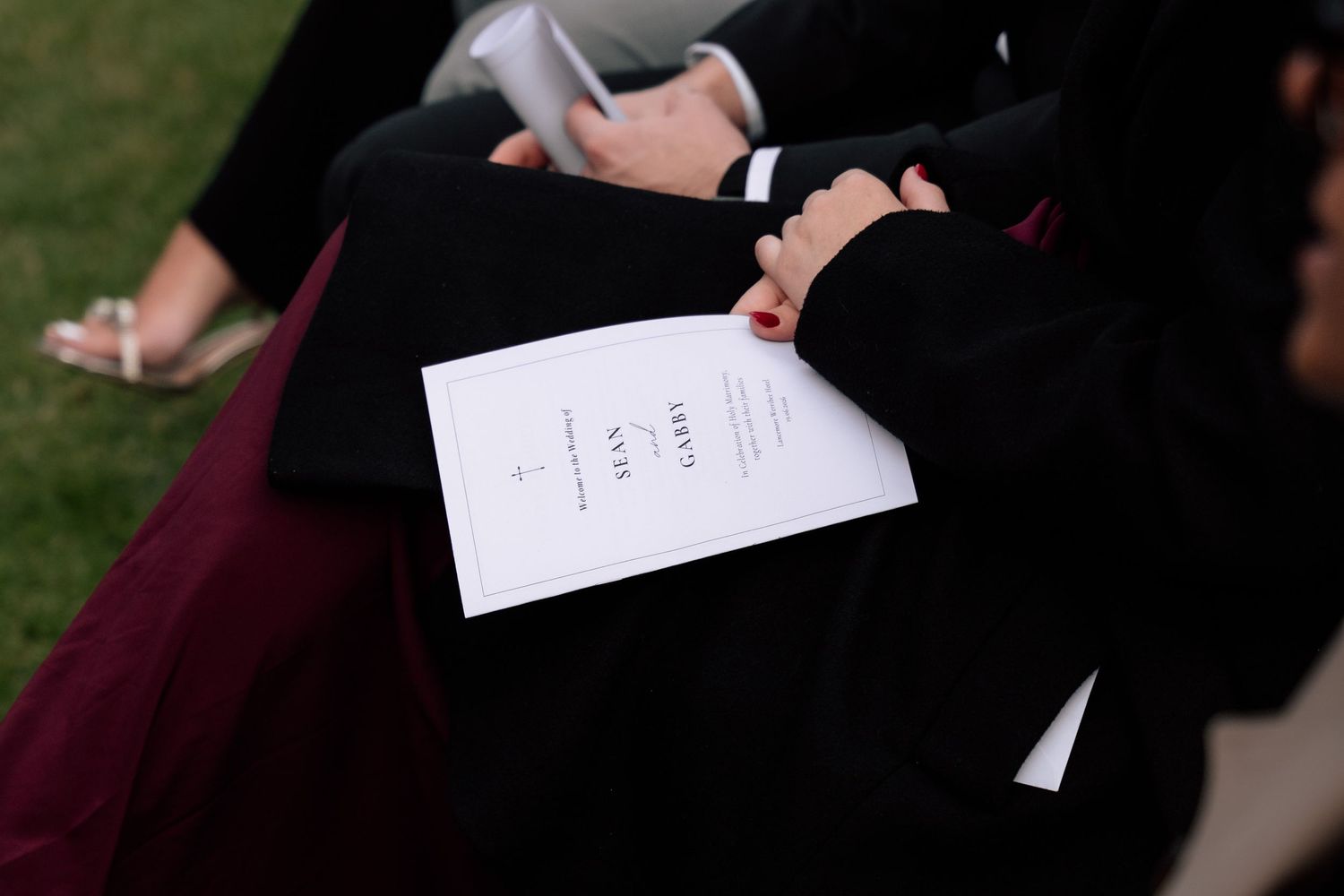 Person in black attire holding a formal white wedding program during an outdoor ceremony.