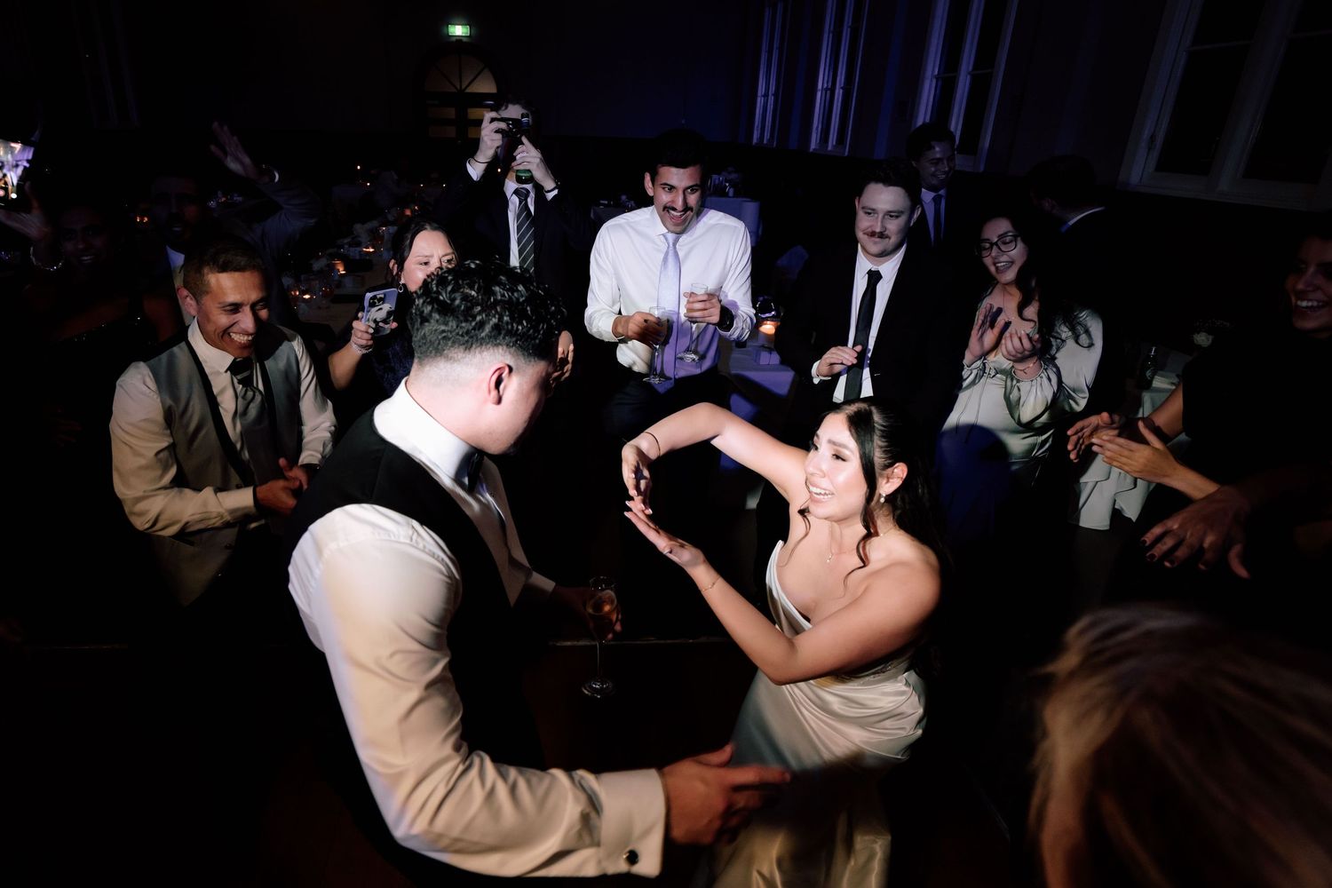 Bride and groom share dance surrounded by celebrating guests on dimly lit wedding reception dance floor.