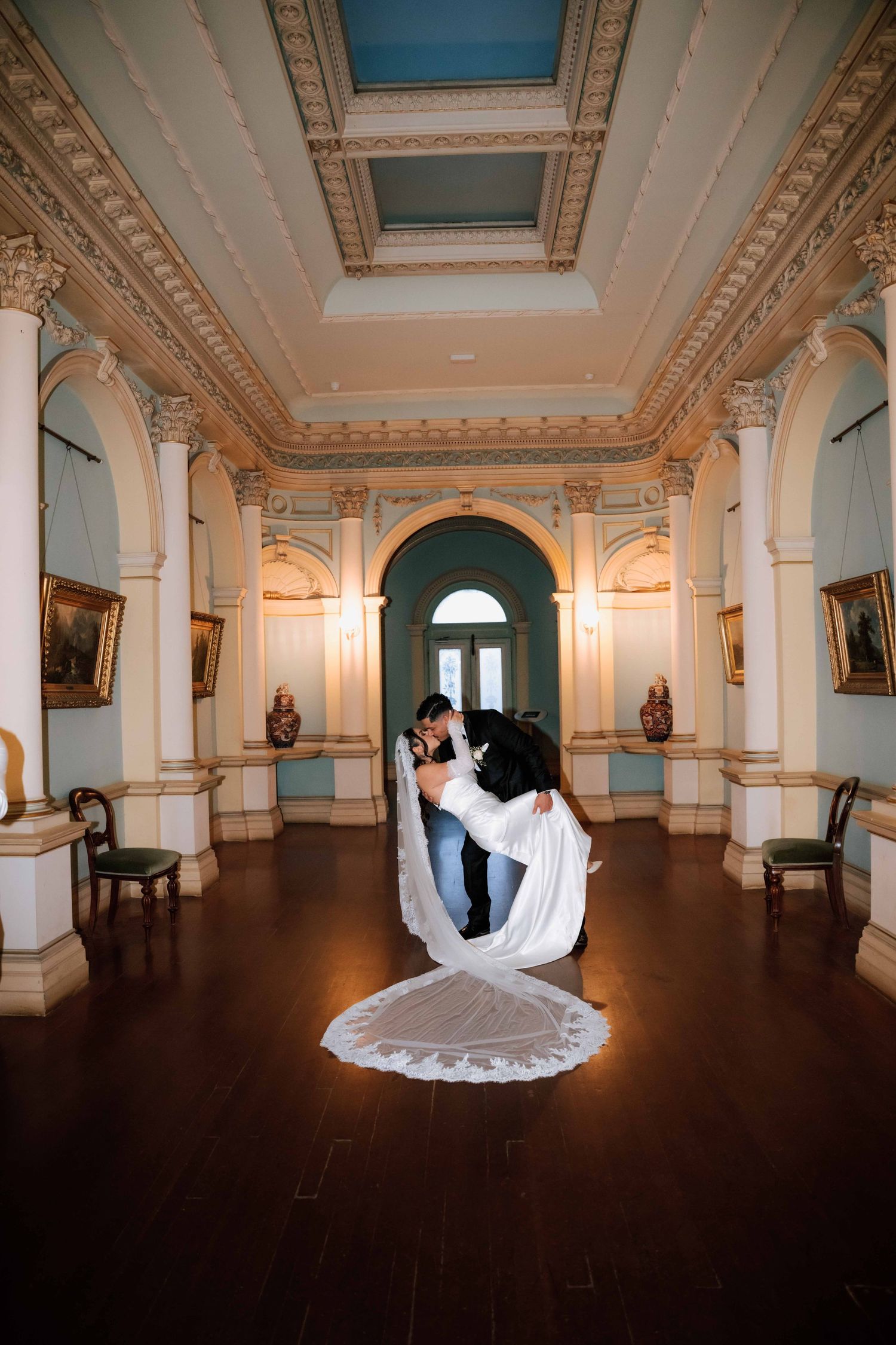 Groom dips bride in a grand ornate ballroom with high ceilings, columns, and elegant decor.