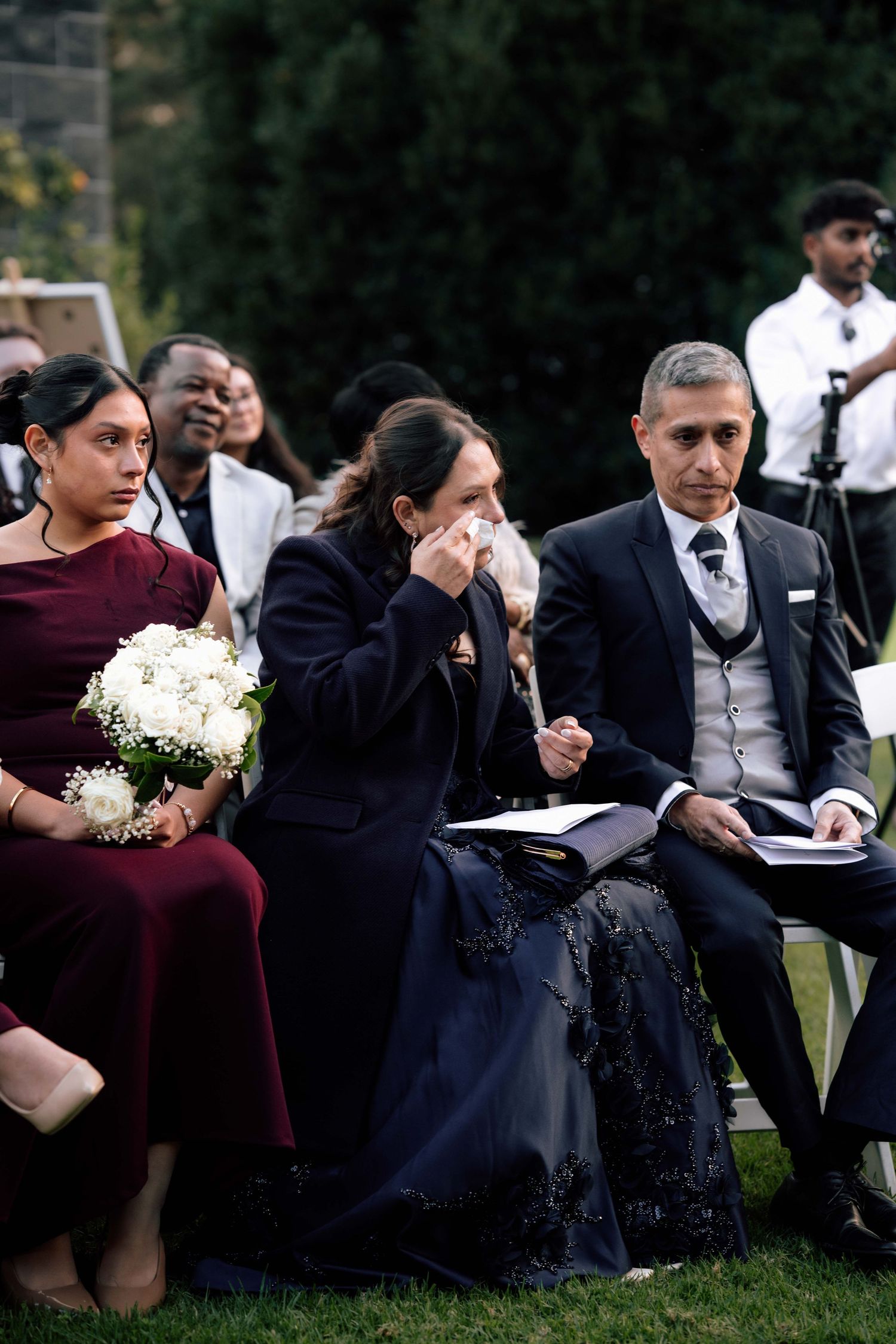 Tearful wedding guests seated outdoors, woman dabbing eyes while man in suit reads ceremony program.