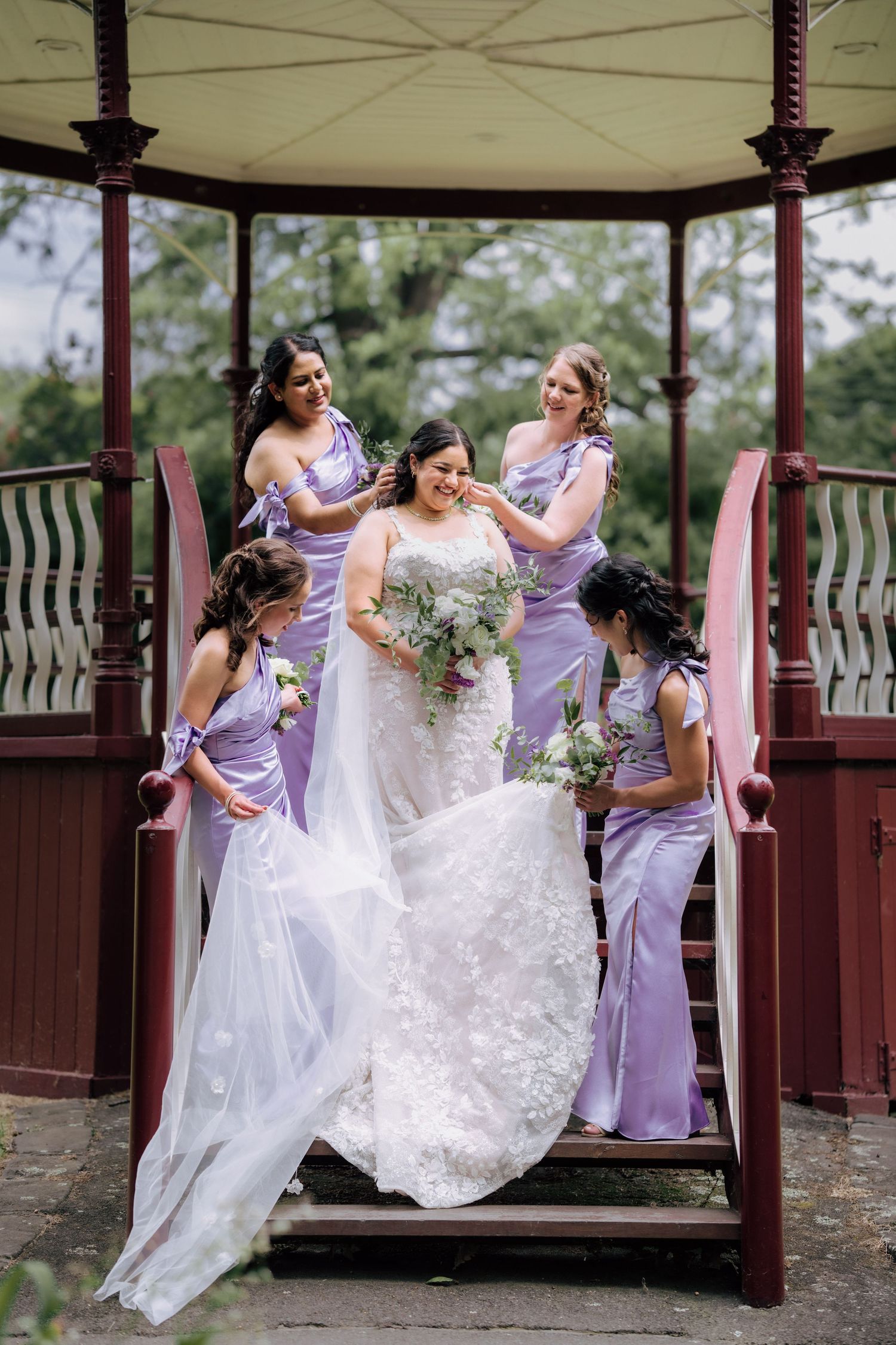 Bride surrounded by four bridesmaids in lavender dresses at red wooden gazebo.