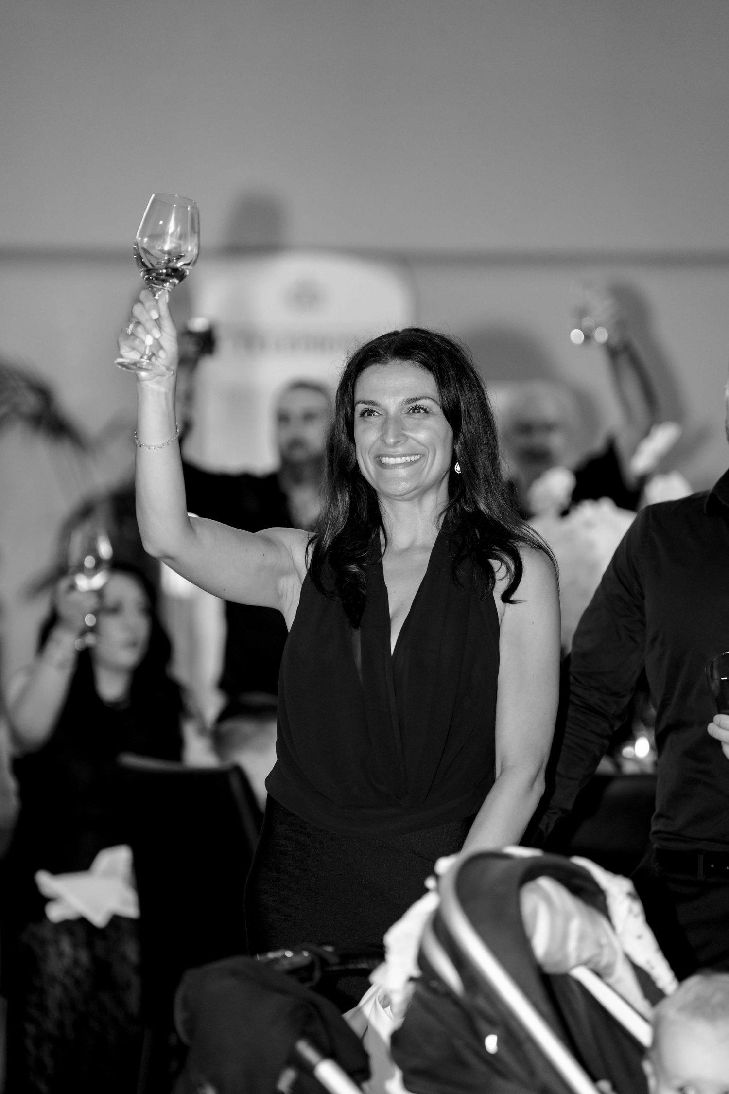 A smiling woman in a black dress raising a wine glass during a toast at a wedding reception.