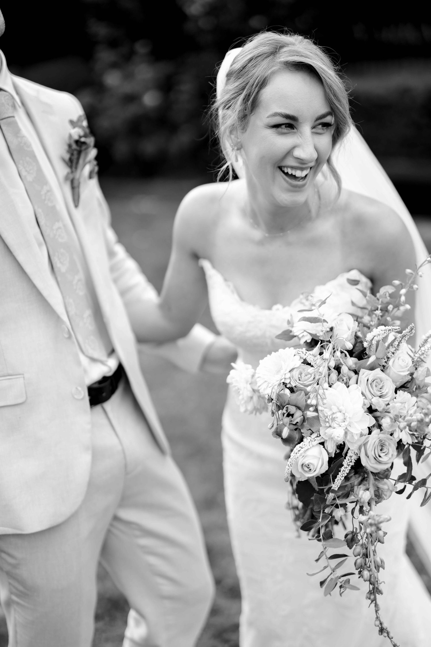 Laughing bride in strapless lace gown holding floral bouquet walks with groom in tan suit at outdoor wedding.