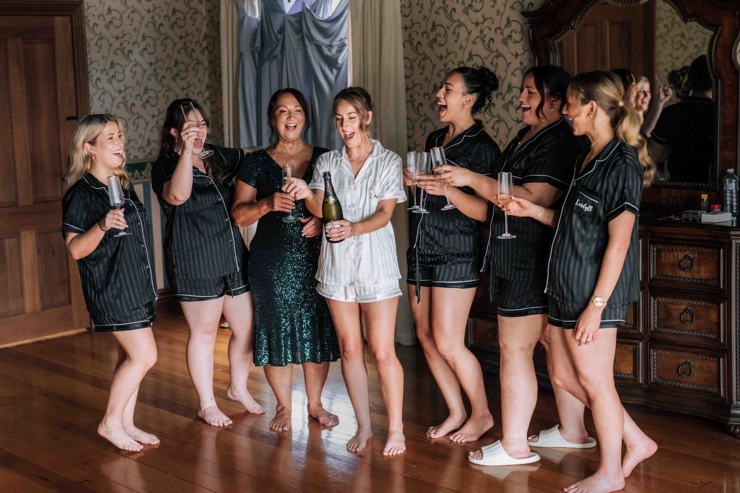 Bride and bridesmaids in matching black satin pajamas celebrating with champagne glasses indoors.
