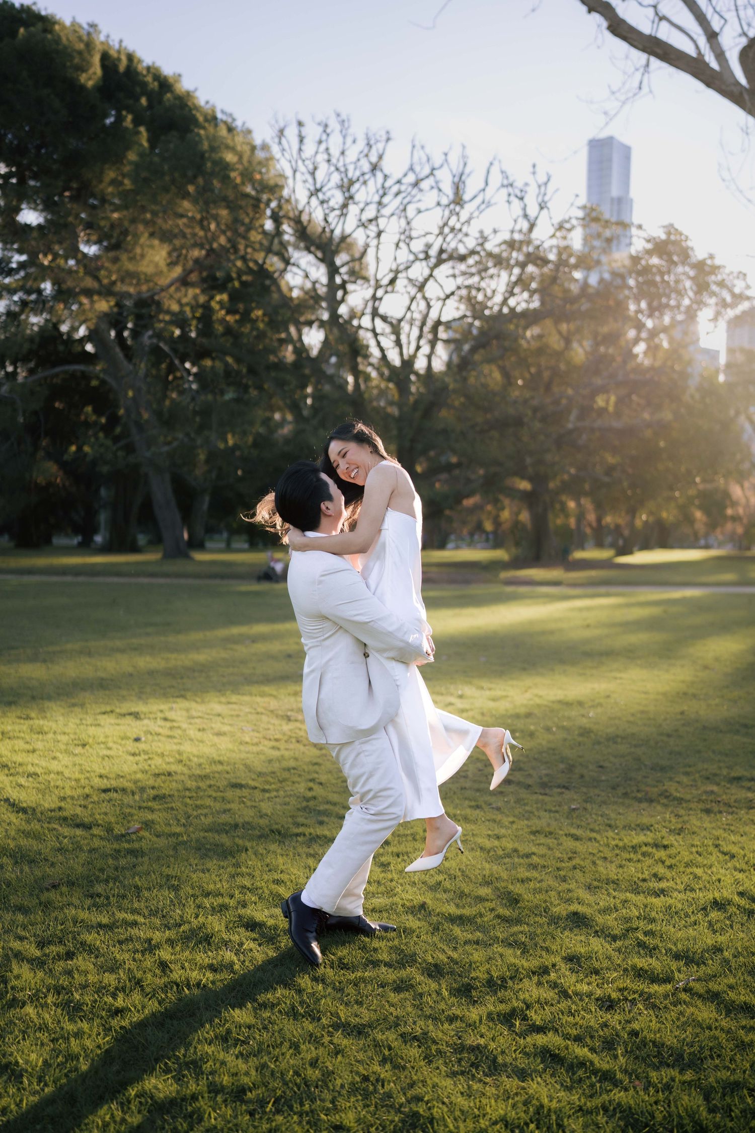 Groom lifting bride in white wedding attire in a sunlit park with lush green grass and trees.