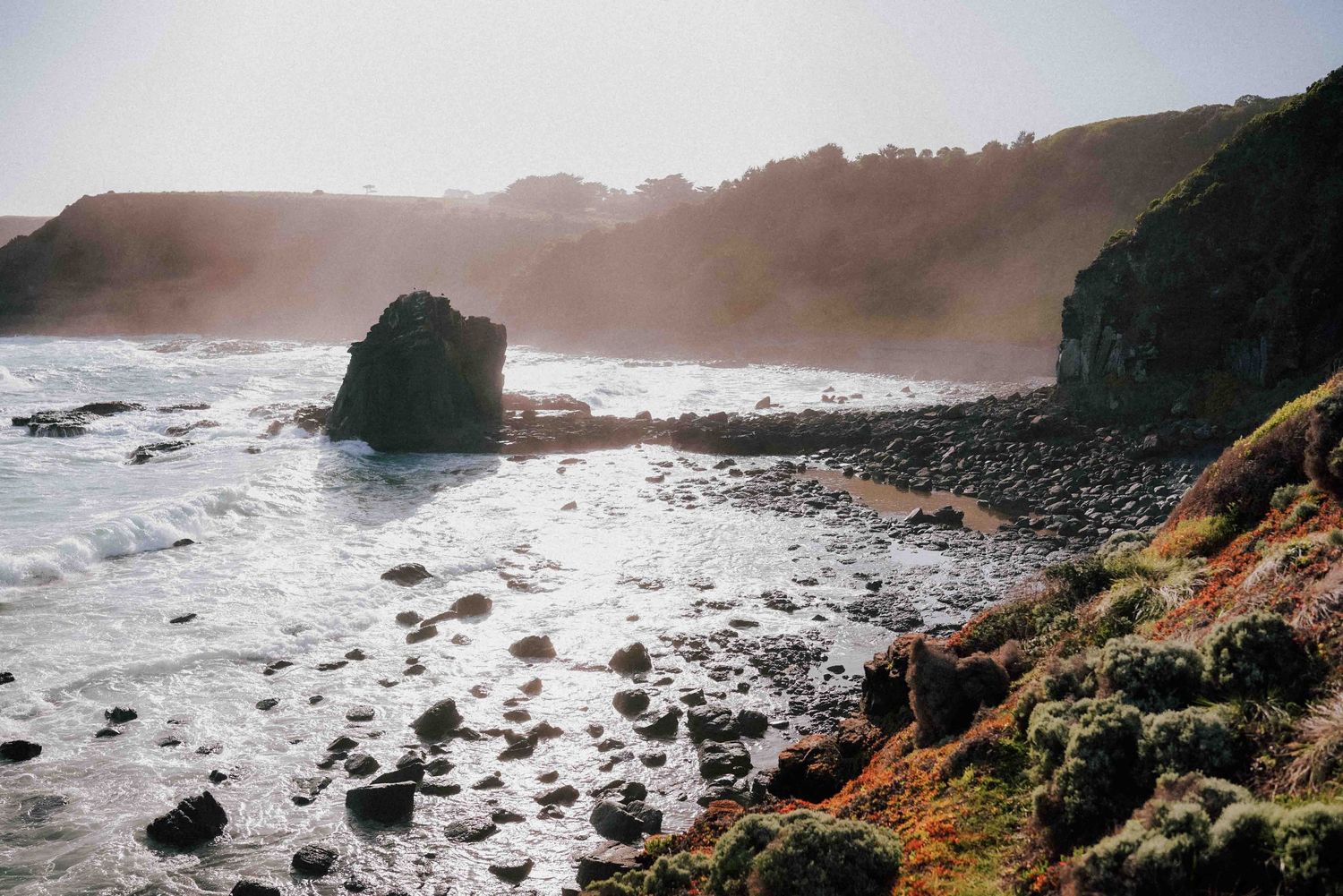 Dramatic coastal landscape with rocky shoreline, sea mist, and lush green cliffs.