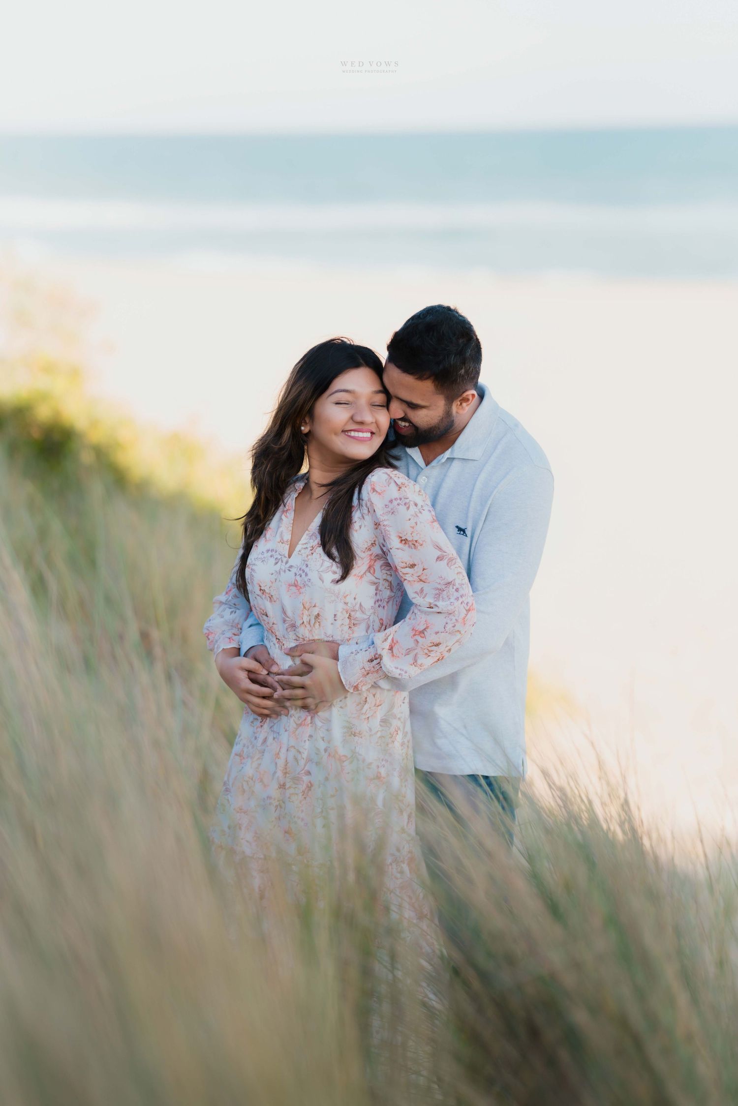 Couple cuddling in seaside dune grass, woman in floral dress smiling as partner holds her from behind.