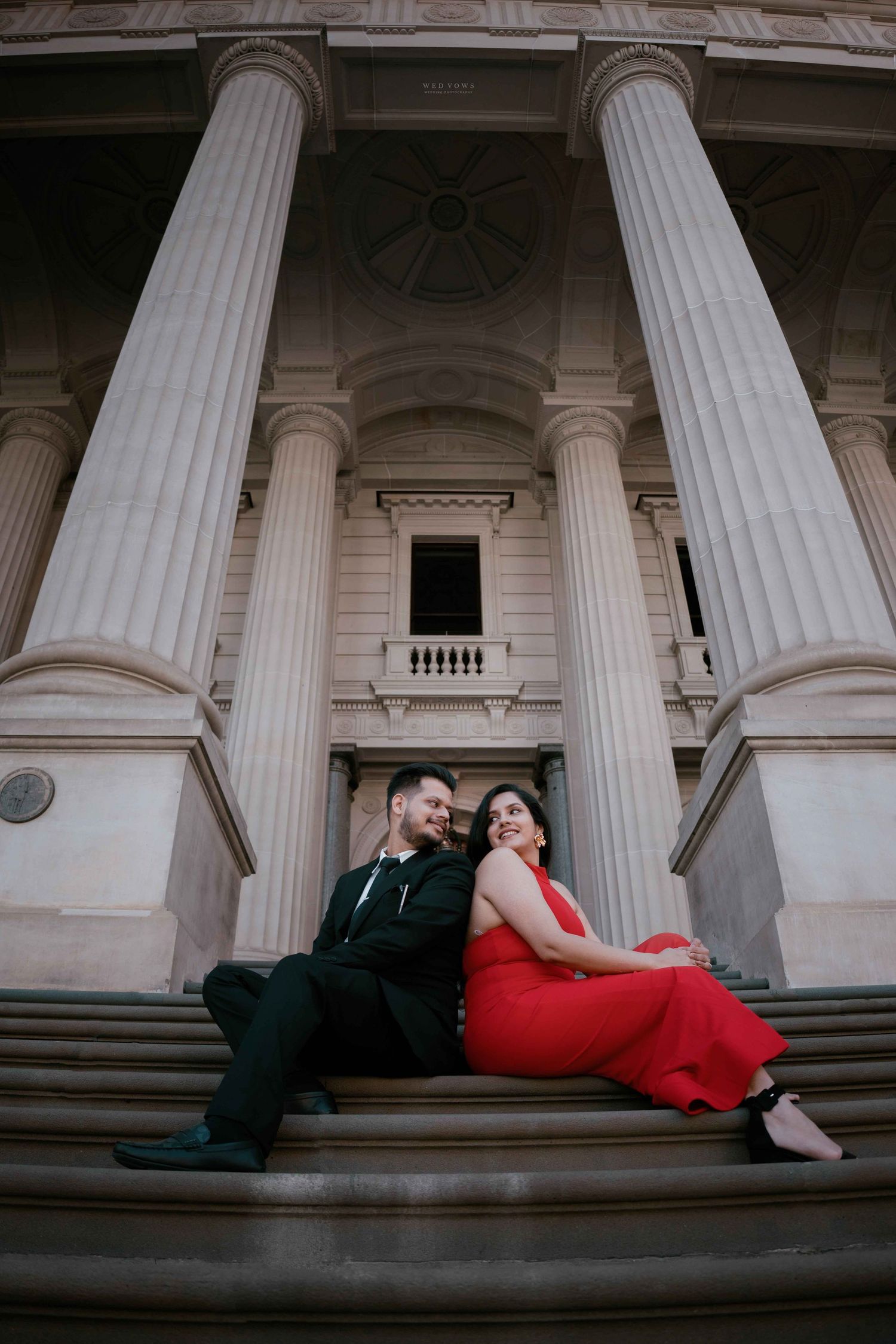 Couple sitting back-to-back on steps of grand columned building in elegant red and black outfits.