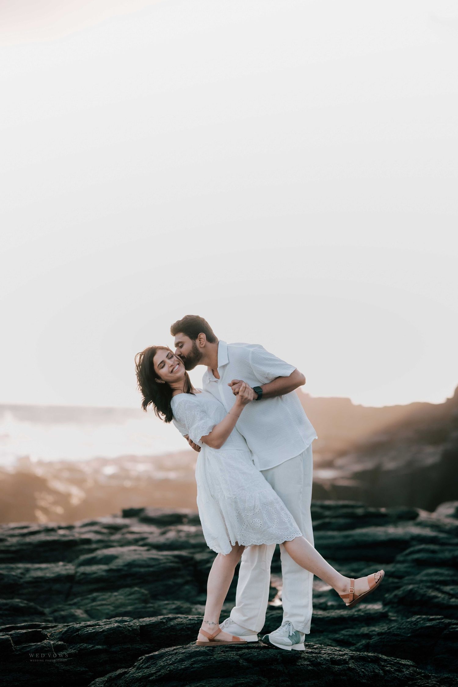 Man in white dipping woman in white dress while kissing her on dark volcanic rocks at sunset.