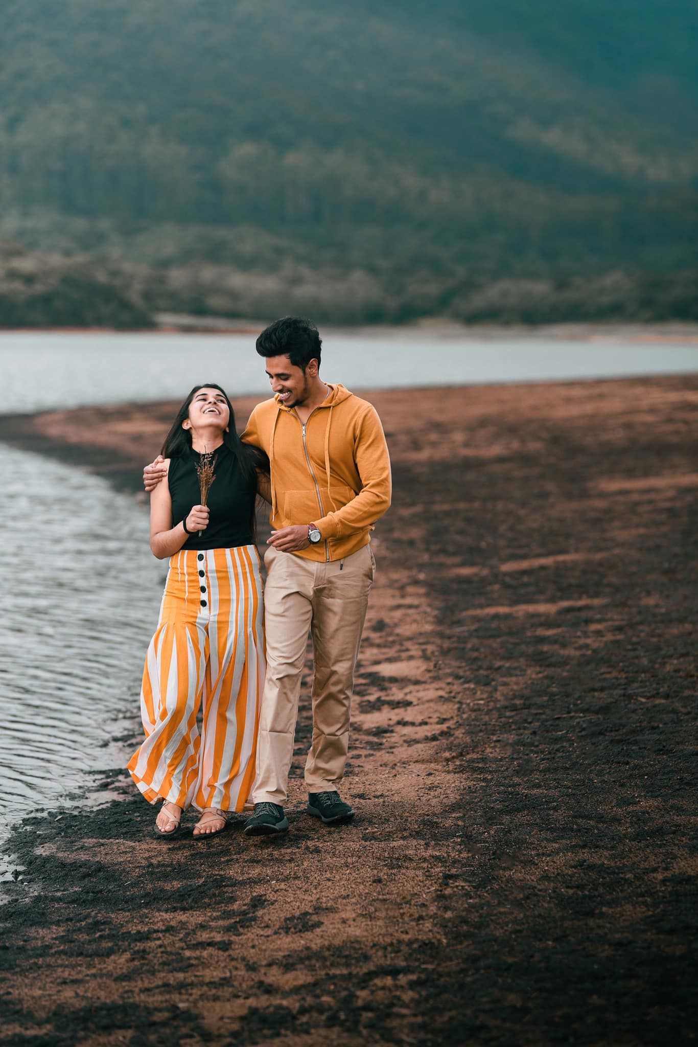 Couple walking together along a lakeside path with mountains in the background.