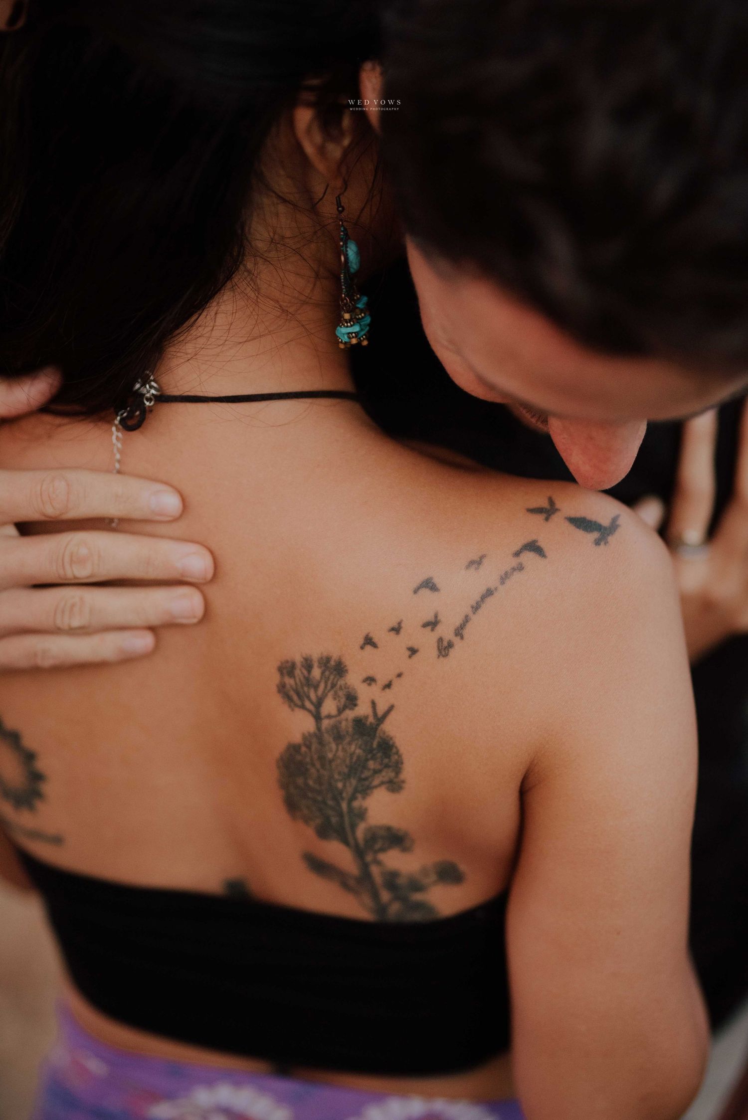 Close-up of a womans back tattoo featuring a tree with birds in flight, with a mans hand on her shoulder.