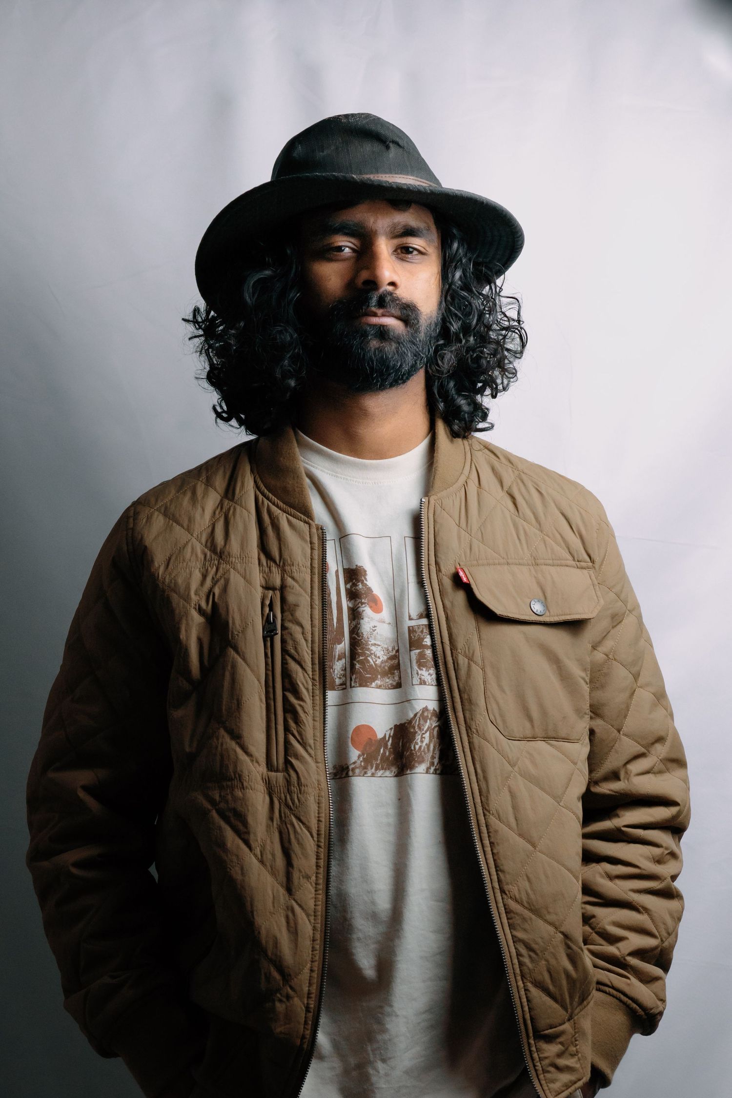 Stylish man with curly hair and beard wearing a hat and quilted jacket in a studio portrait.