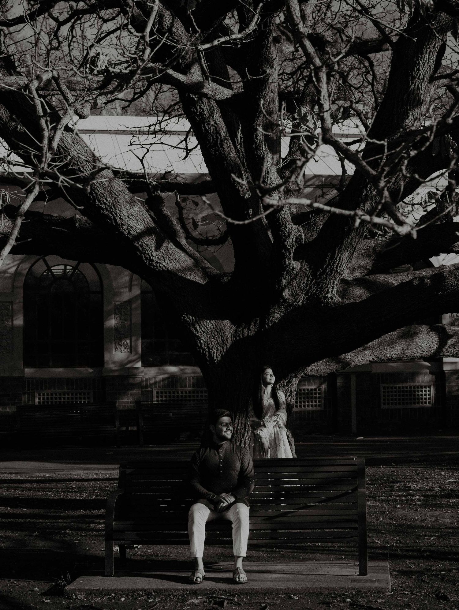 Black and white portrait of a couple beneath a sprawling bare tree, man seated on bench, woman standing.