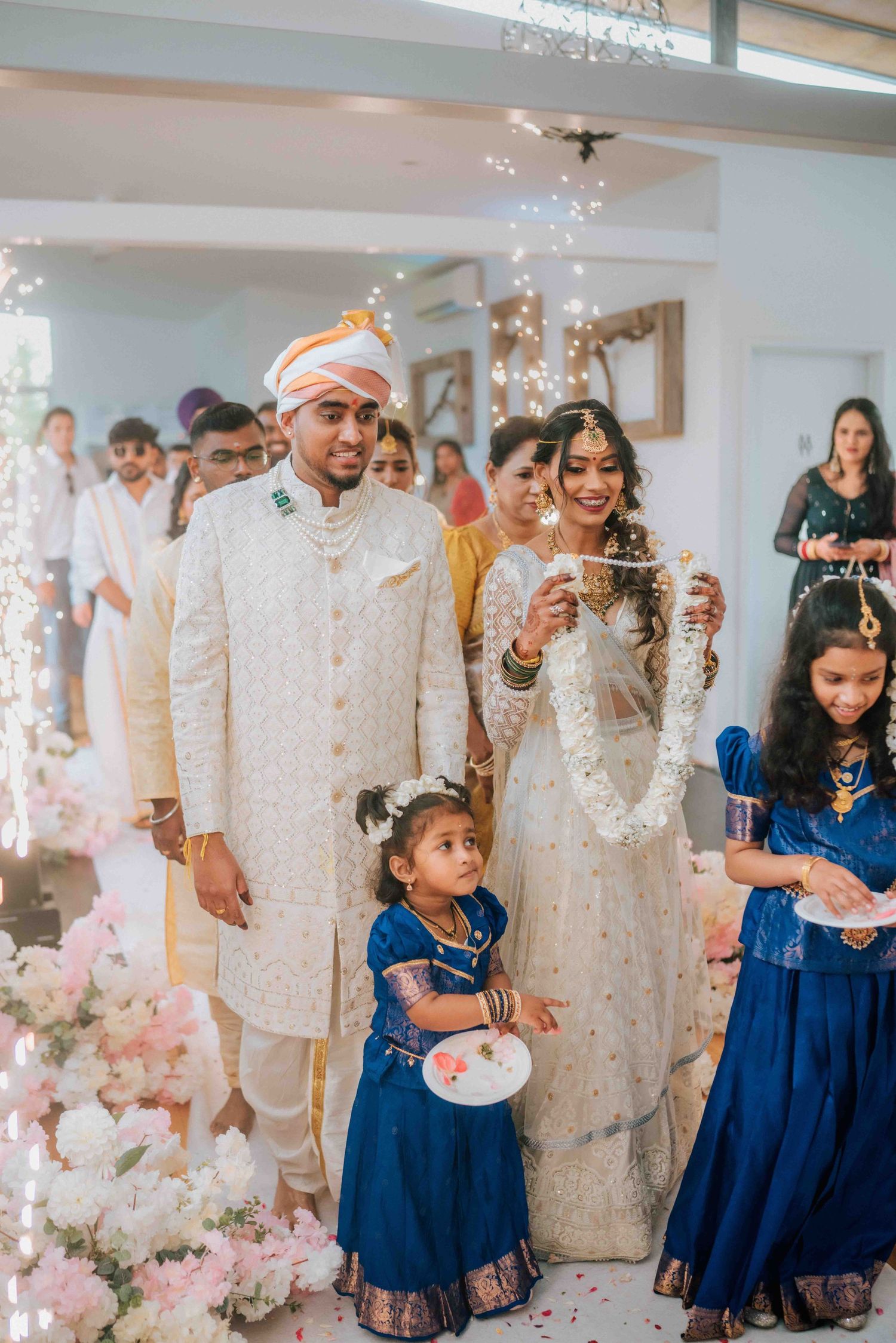 Indian groom in white sherwani and bride in elegant attire walk with children at wedding ceremony.