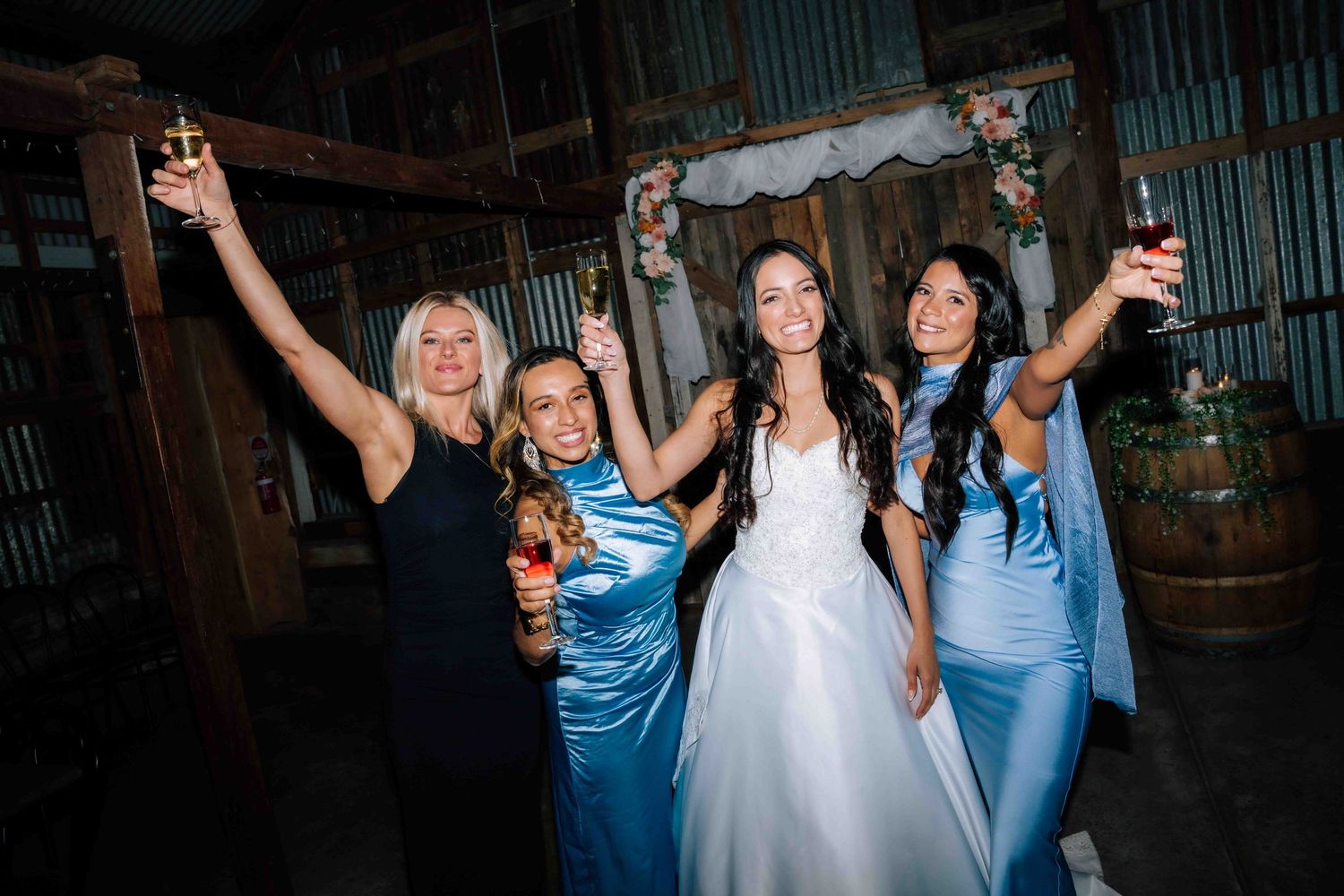 Bride in white gown celebrates with three women in blue dresses raising glasses at wedding.
