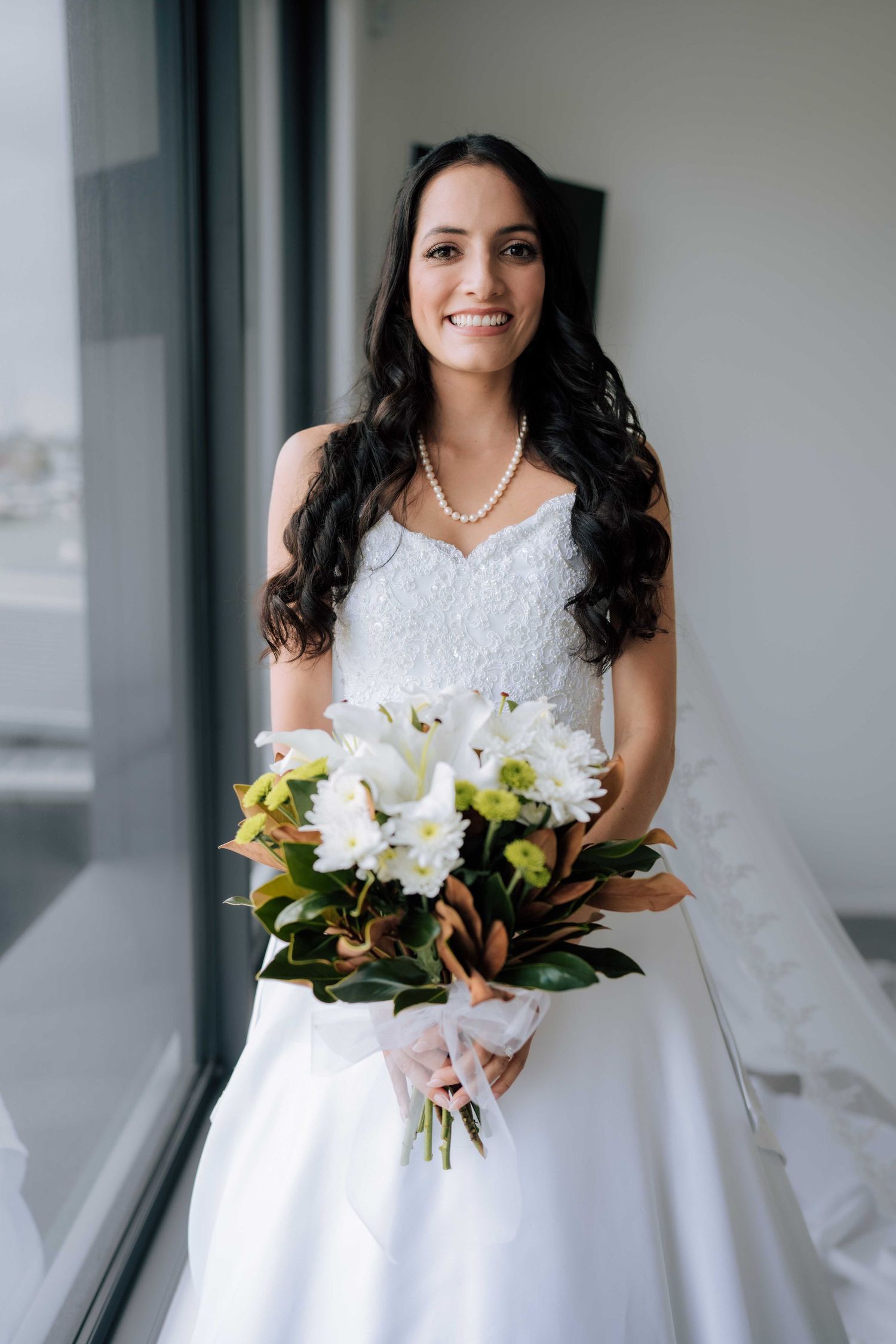Smiling bride with dark curly hair holds white floral bouquet wearing pearl necklace by window.