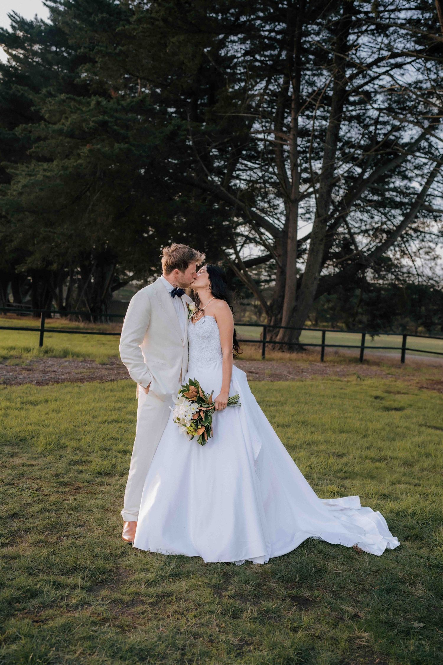 Bride and groom share a romantic kiss in a lush green outdoor setting, bride holding a beautiful bouquet.