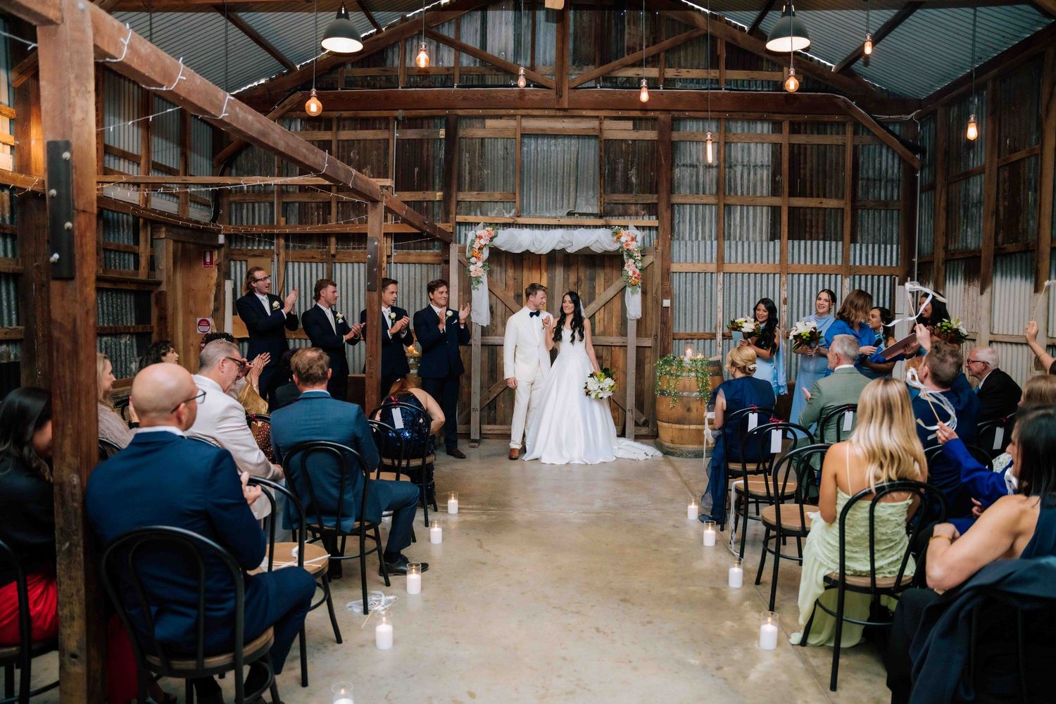Rustic barn wedding ceremony with couple at floral arch altar, guests seated in black chairs with candles lining the aisle.