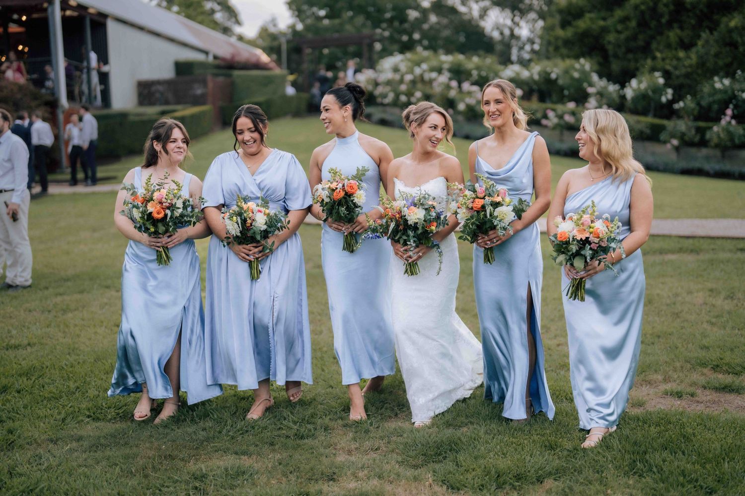 Bride in white gown walks with five bridesmaids in light blue satin dresses holding colorful bouquets on green lawn.