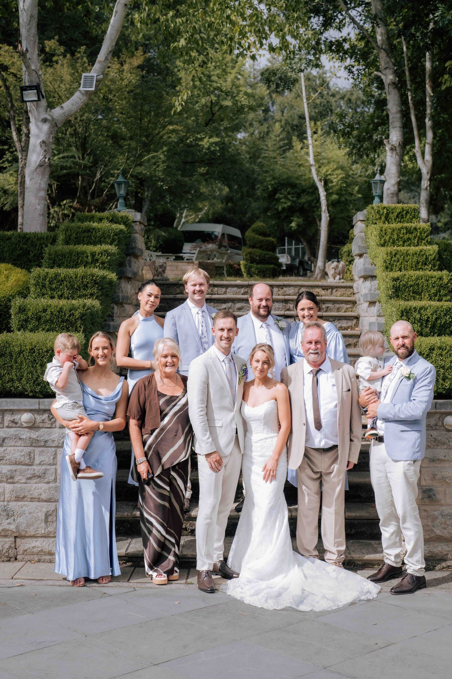Bride and groom celebrate with entire family group on stone steps at garden wedding.