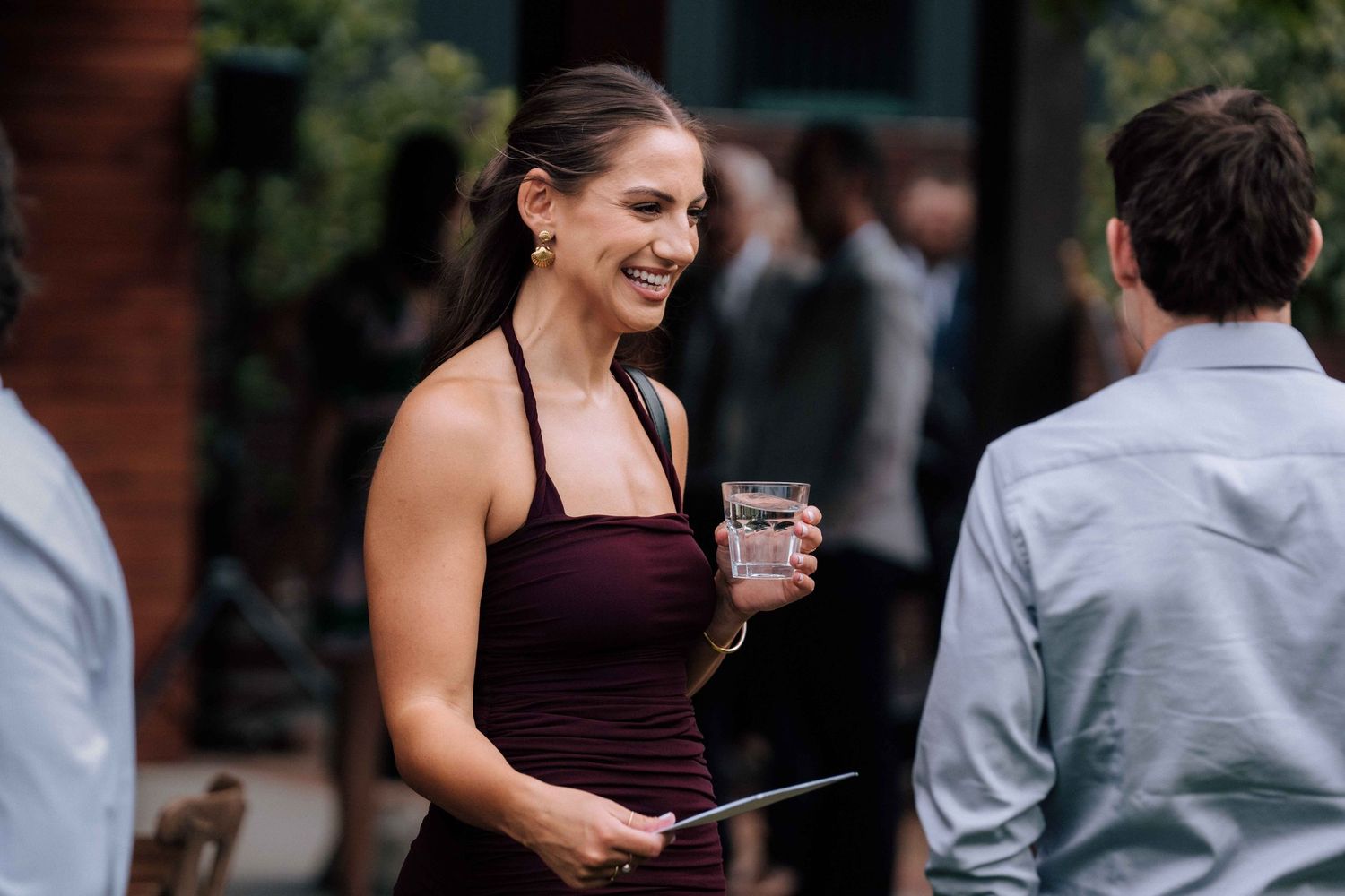 Smiling woman in a burgundy dress holding a drink while socializing at an outdoor wedding reception.