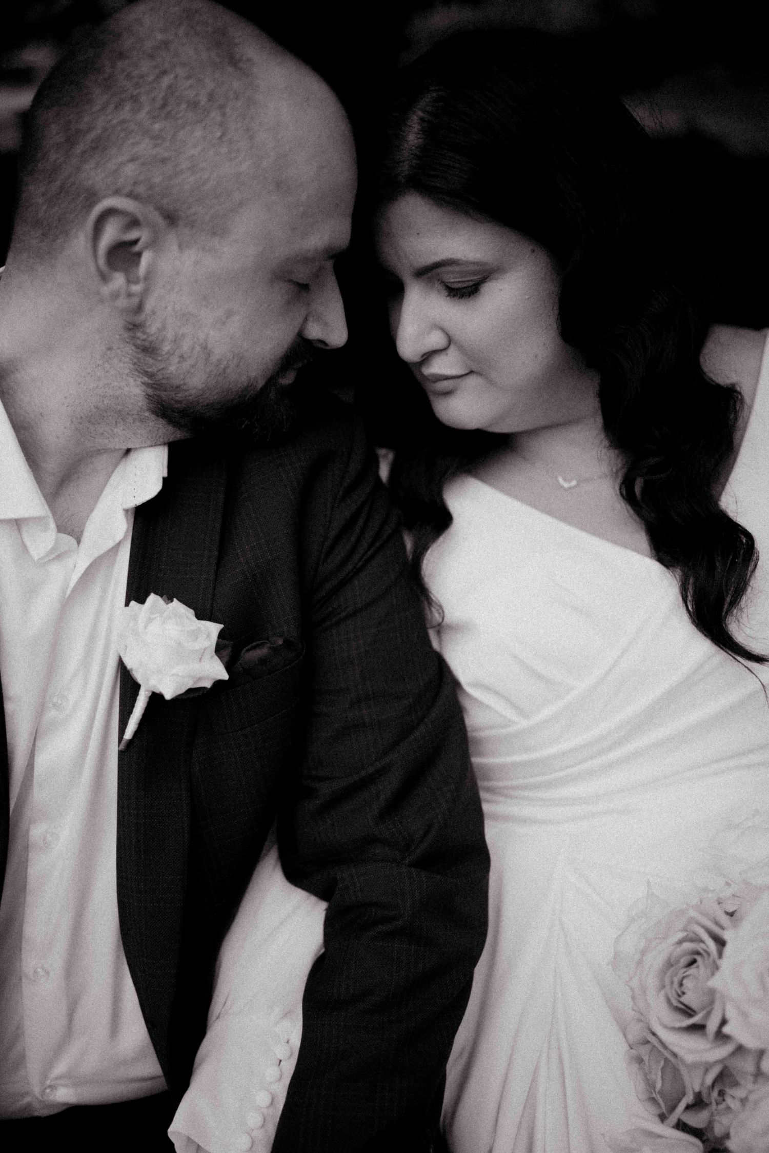 Black and white photo of bride and groom sharing an intimate moment, groom in suit with boutonniere, bride holding roses.