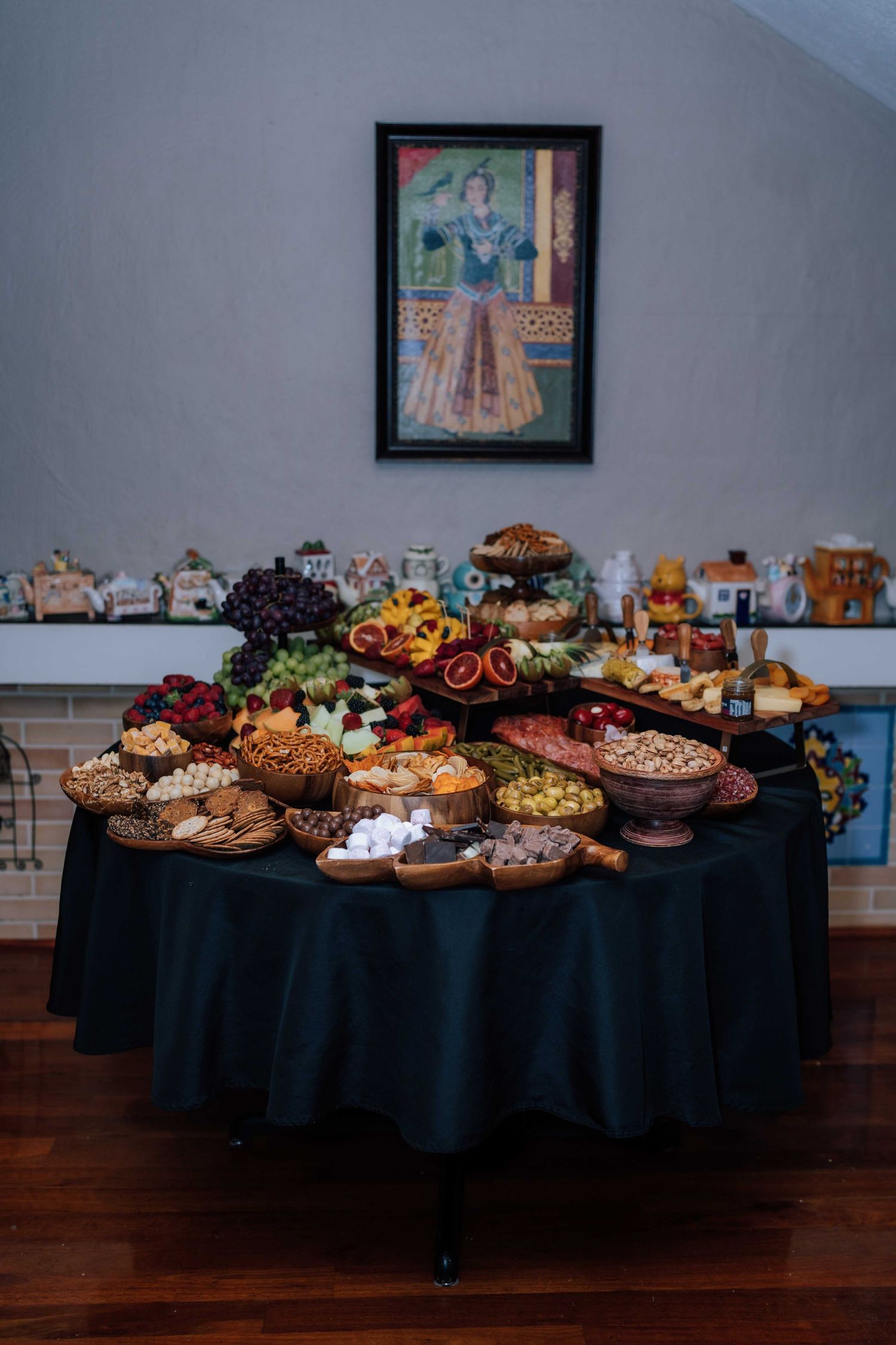 Elegant wedding grazing table with fruits, nuts, and snacks on black tablecloth beneath framed artwork.