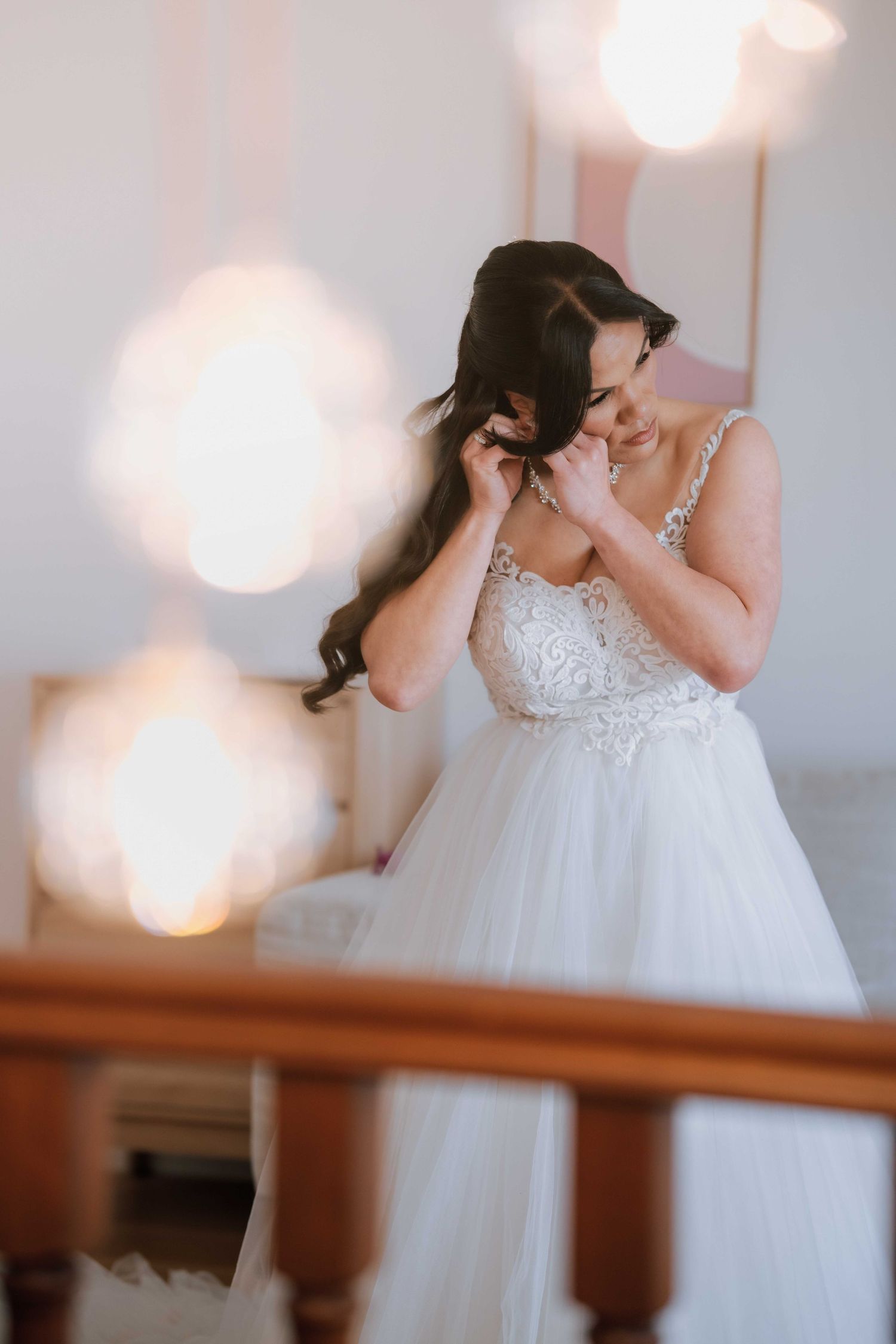 A smiling bride in a white lace tulle gown adjusts her earring near a staircase with bokeh lights.