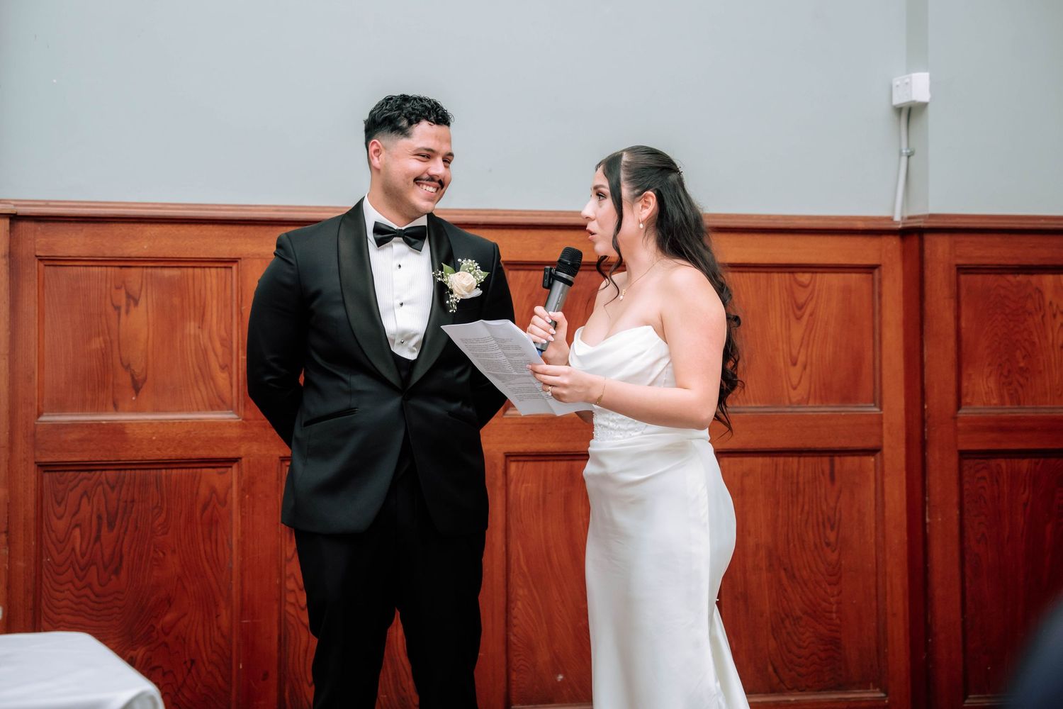 Bride speaking into microphone while groom smiles during wedding reception speeches at head table.