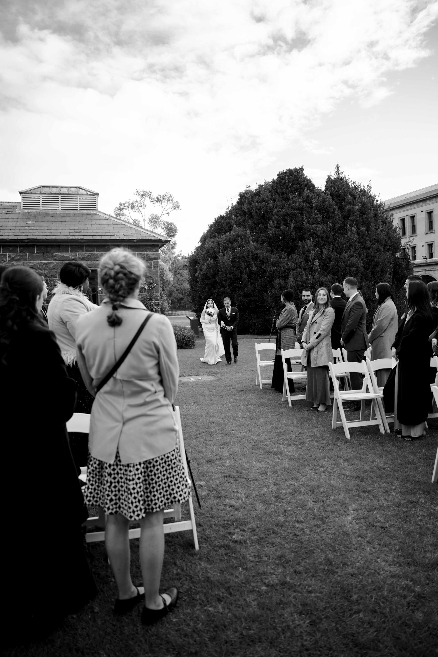Guests stand and watch as a couple exchanges vows during an outdoor wedding ceremony on a lawn.