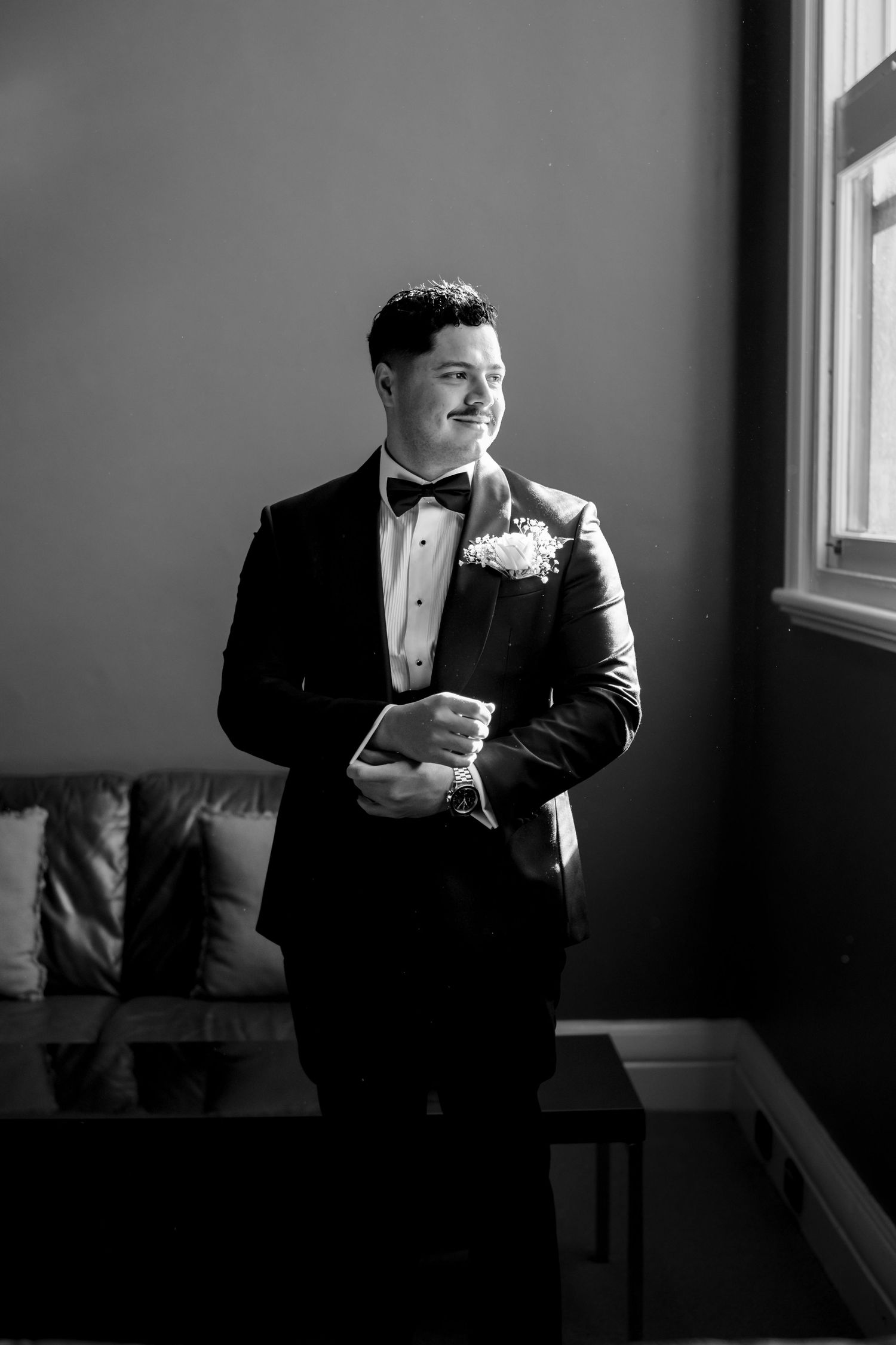 Groom in black tuxedo and bow tie adjusting cufflinks by window in black and white wedding portrait.