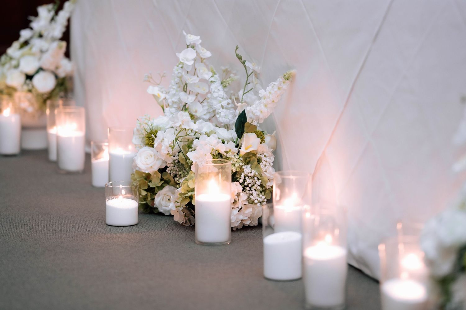 White wedding aisle decor with candles and floral arrangements of white hydrangeas and orchids.