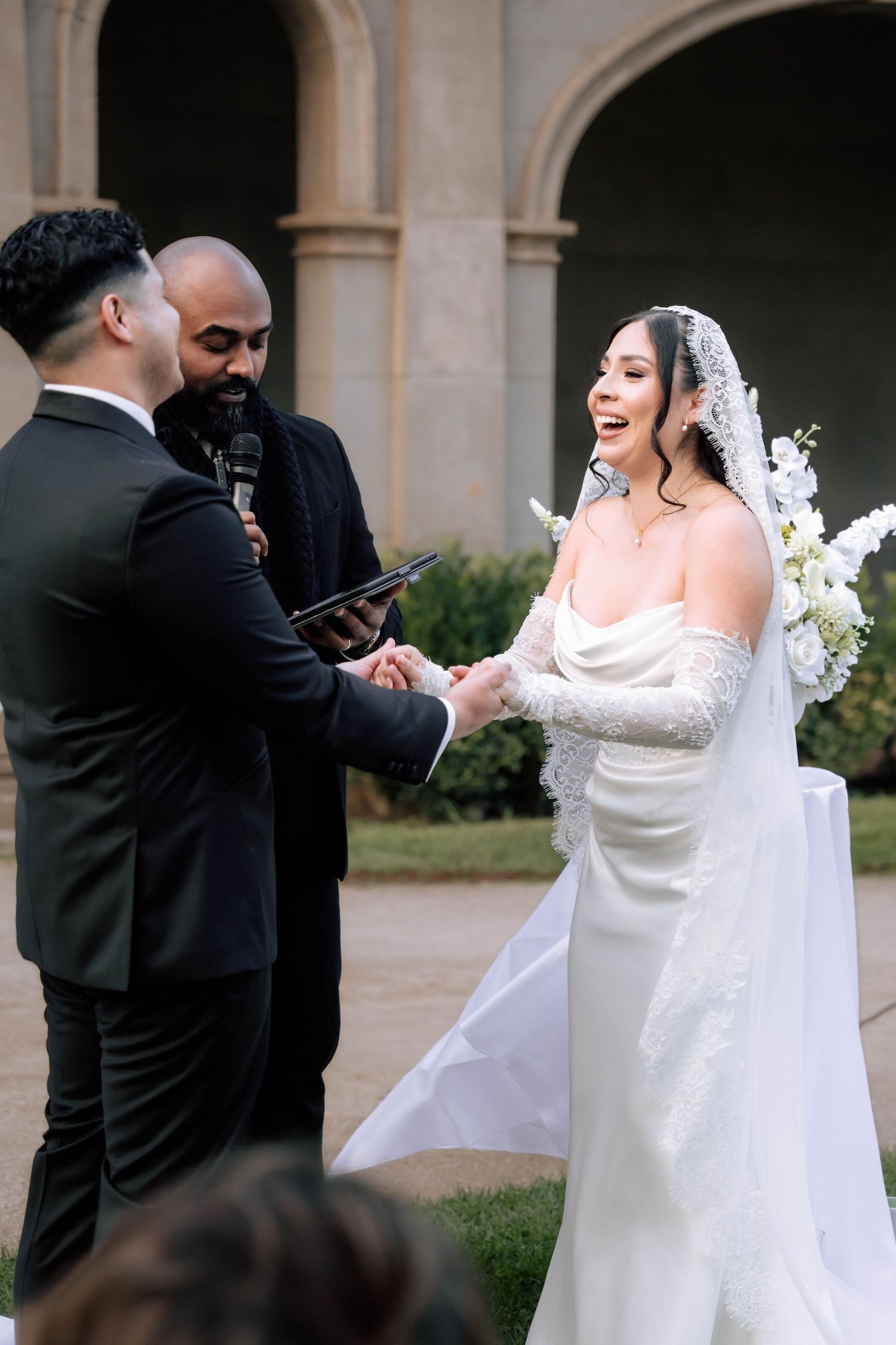 Bride and groom hold hands laughing during outdoor wedding ceremony while officiant reads vows.