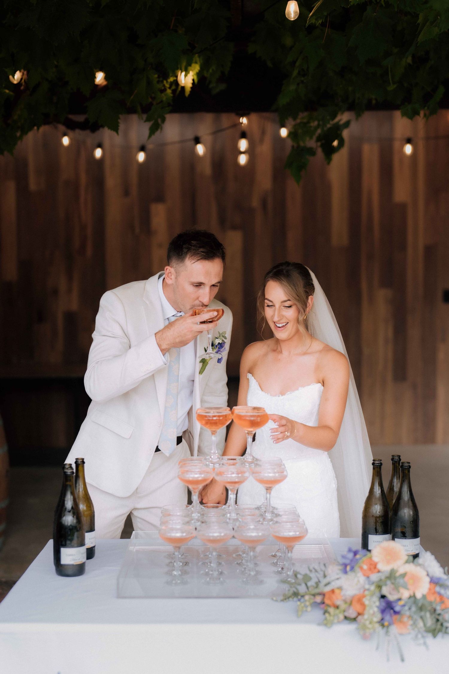 Bride and groom enjoying champagne tower at wedding reception under string lights with wine bottles.