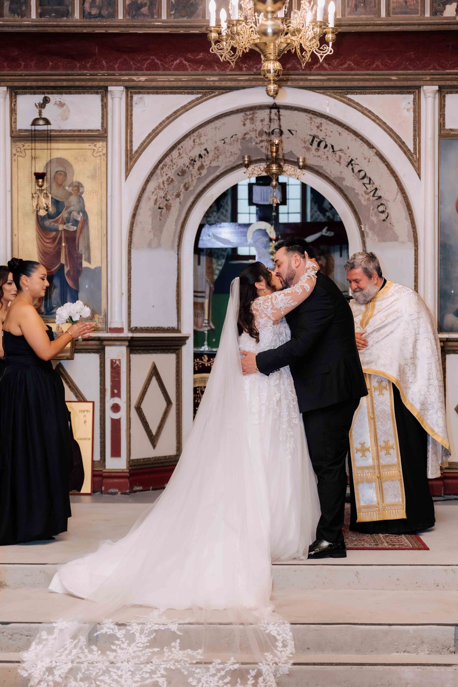 Newlyweds share first kiss at altar in ornate Greek Orthodox church after ceremony.
