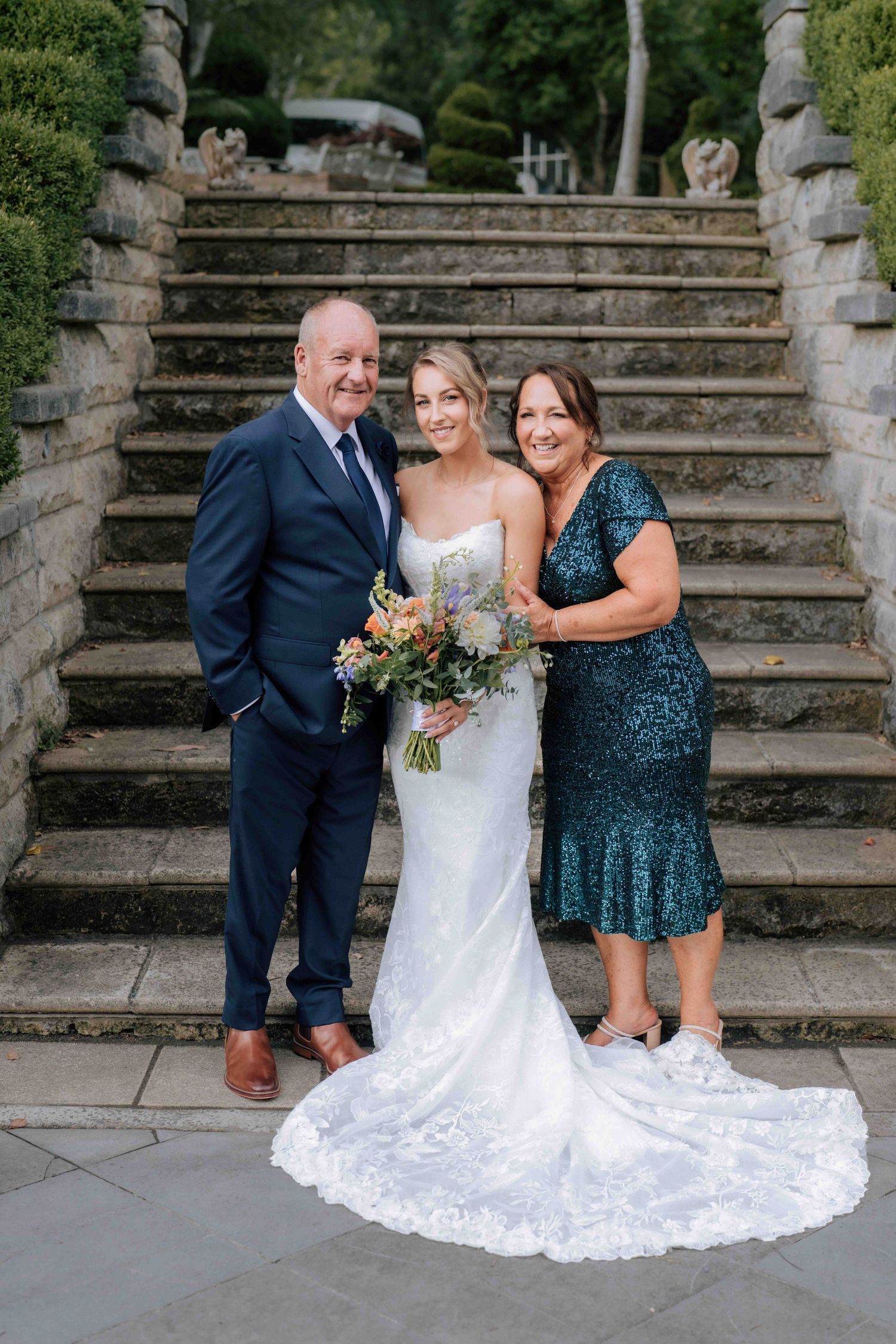 Bride in lace wedding gown holding wildflower bouquet posing with parents on stone steps outdoors.