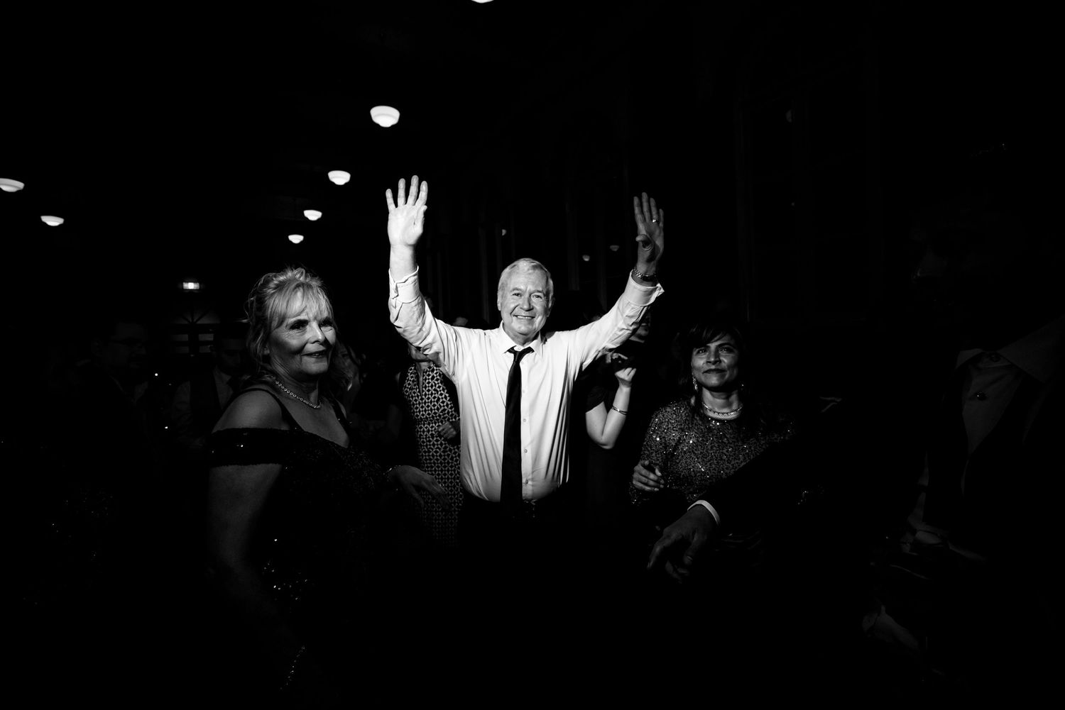 Black and white photo of joyful man dancing with arms raised at wedding reception celebration.