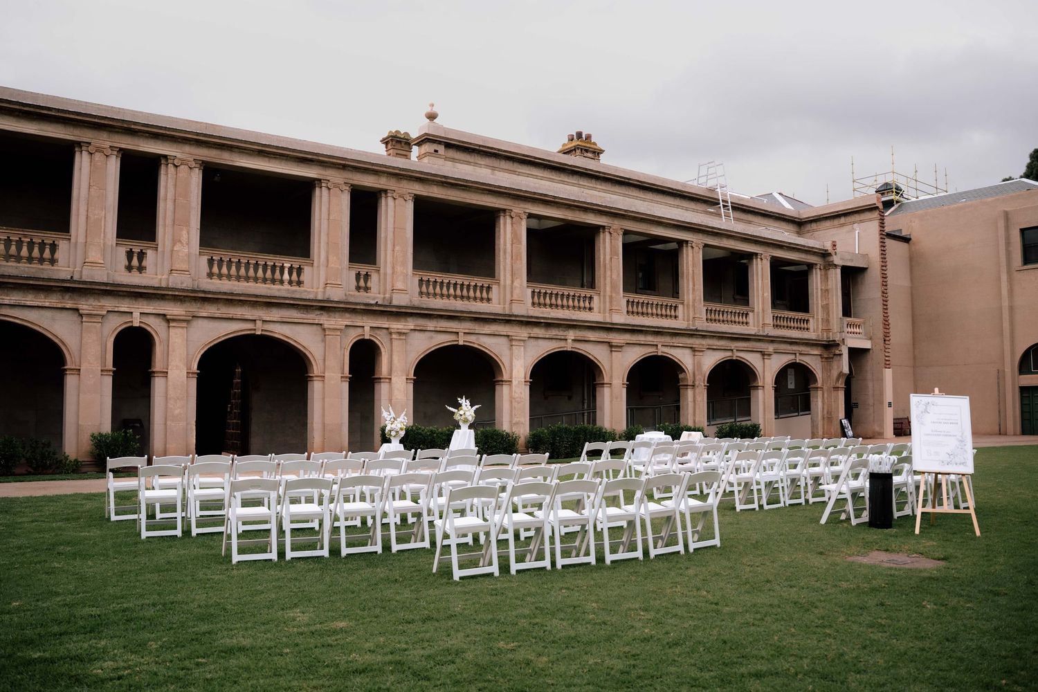 Outdoor wedding ceremony setup with white chairs on green lawn in front of a historic venue.