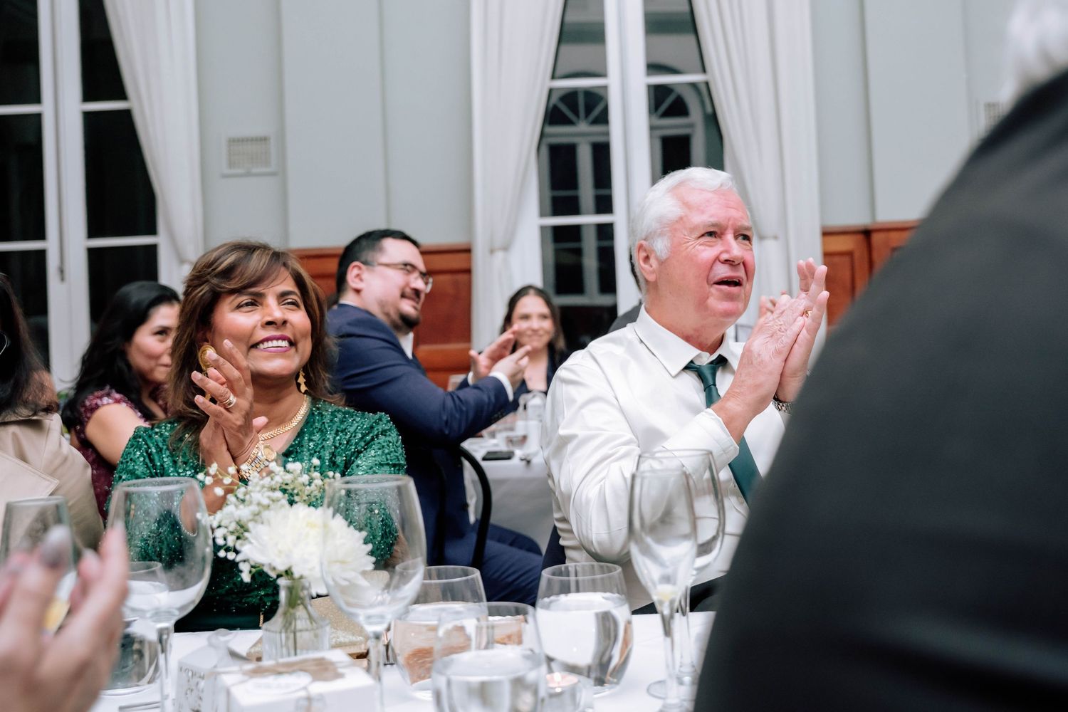 Wedding guests in formal attire clapping and smiling during reception at elegant venue.