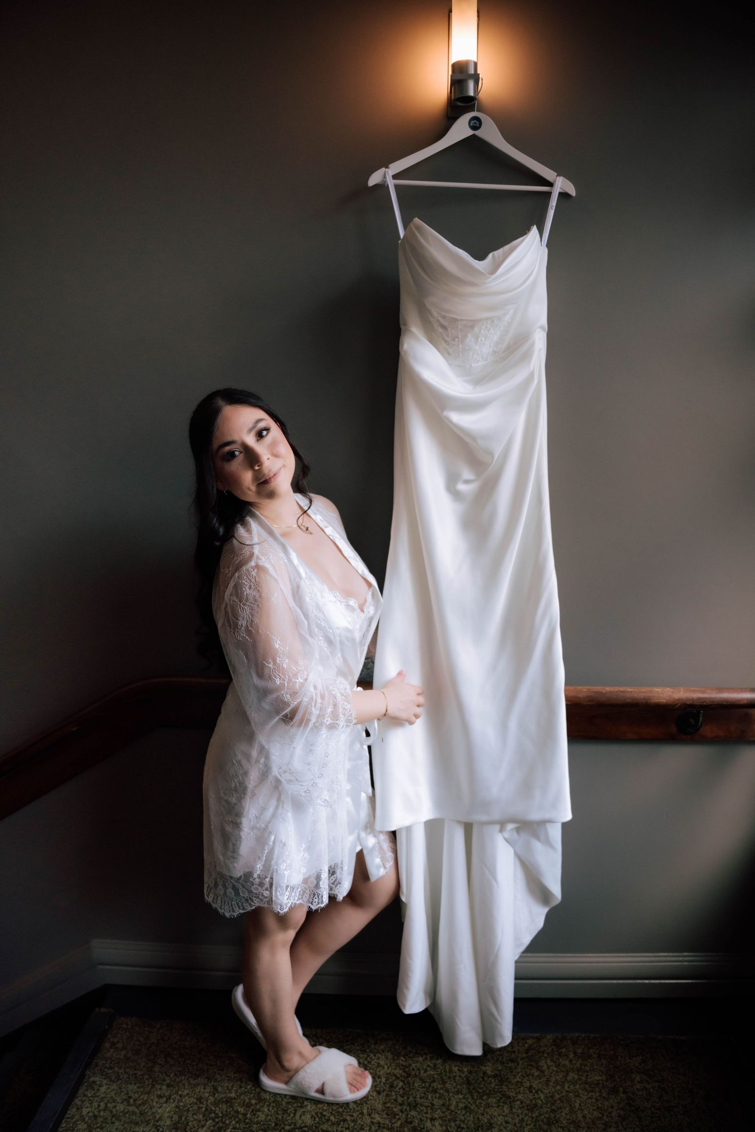 Bride in lace robe standing beside her elegant white strapless wedding gown hanging on a wall.
