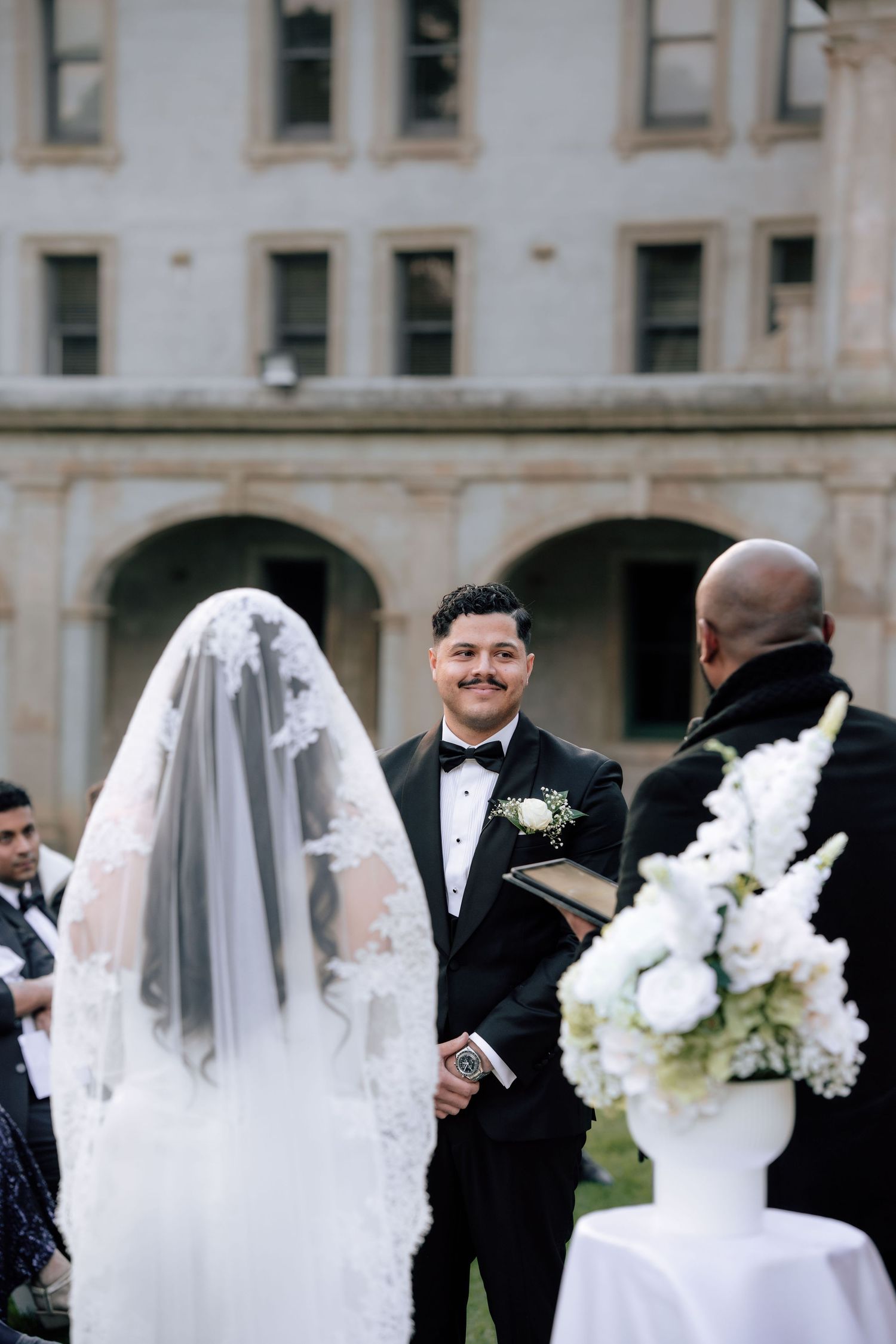 Groom smiles at bride during outdoor wedding ceremony with white floral arrangement and officiant.