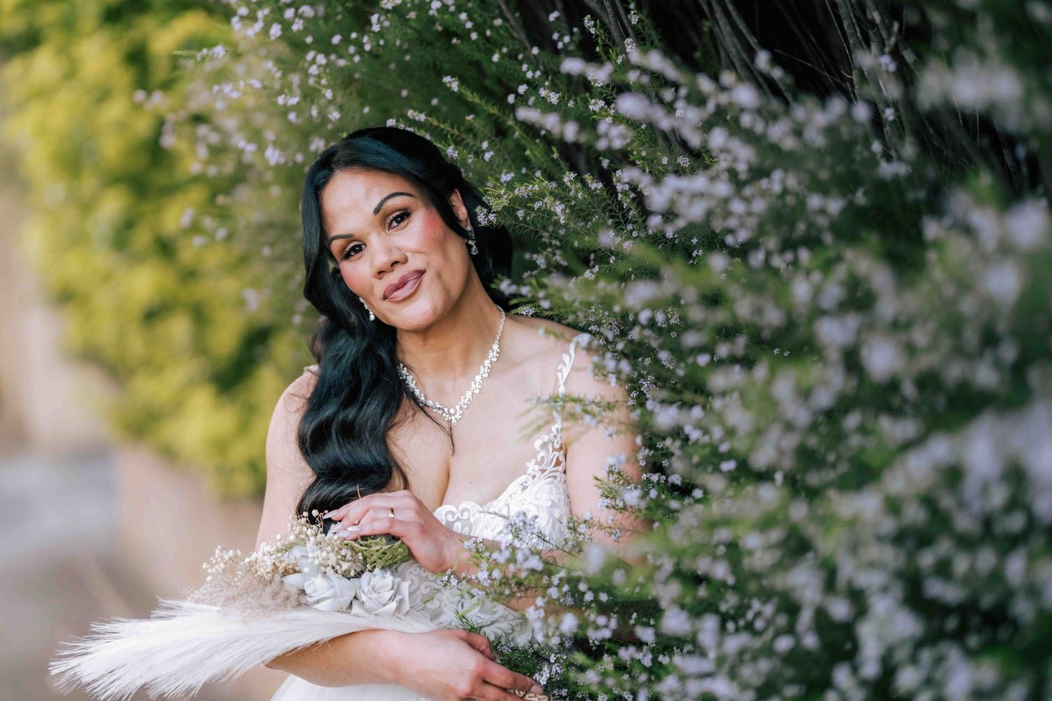 A beautiful bride in a lace wedding dress poses gracefully among delicate white flowering bushes.