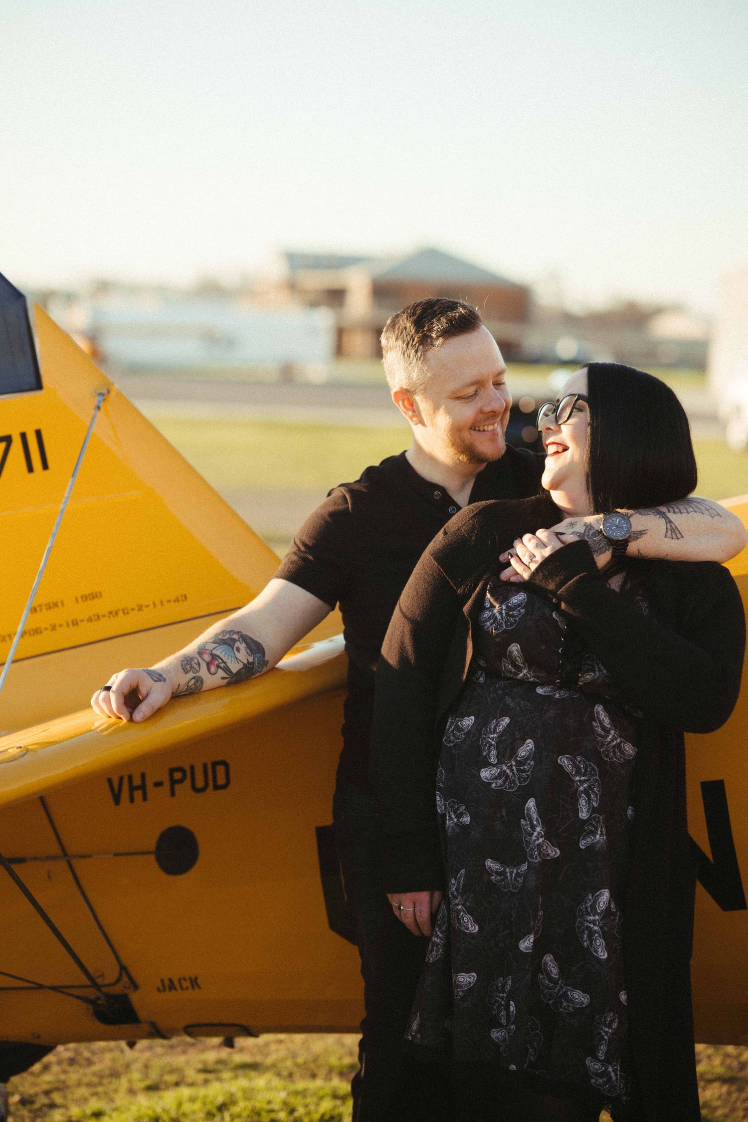 Tattooed couple embracing beside a yellow vintage aircraft registered VH-PUD on an airfield.