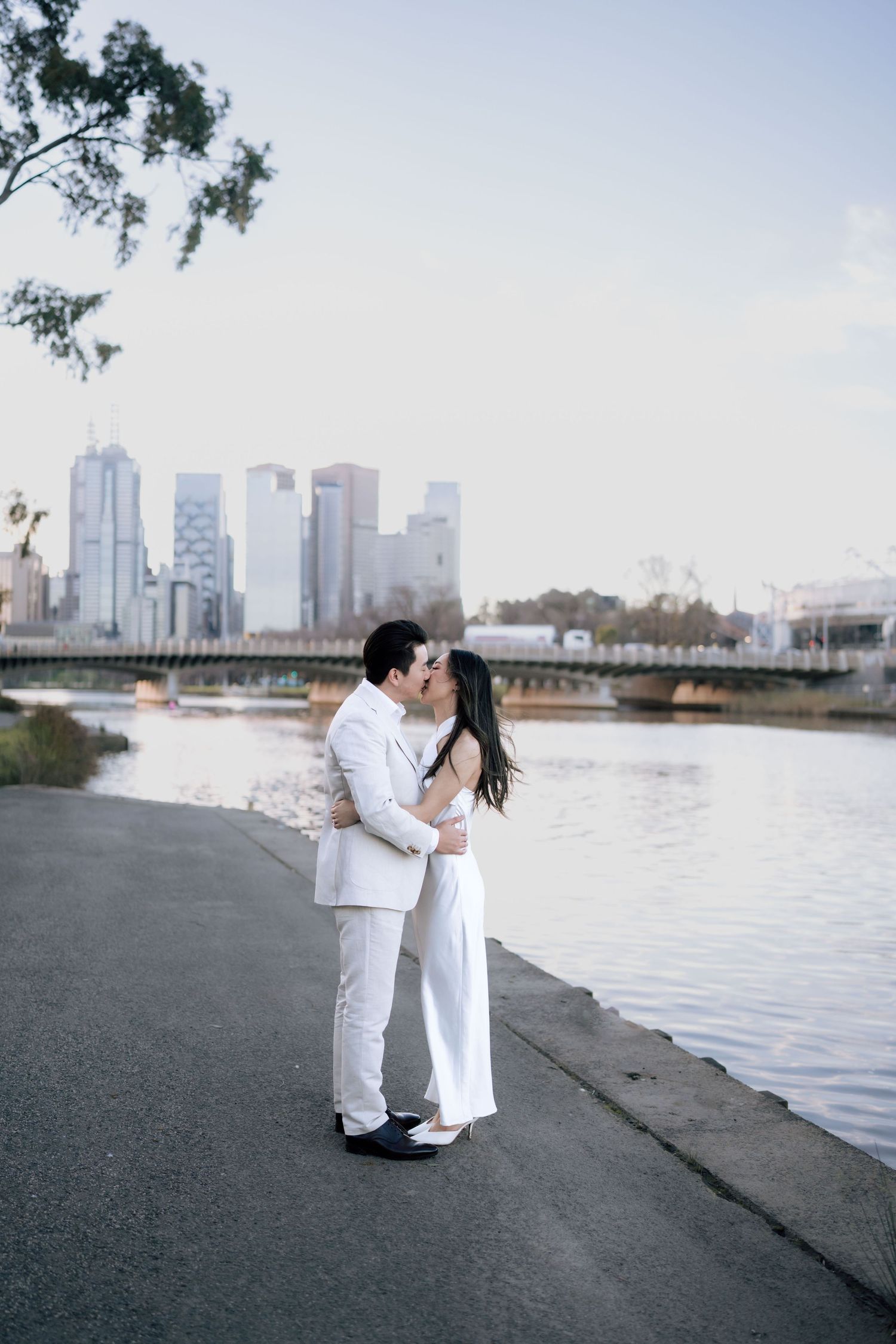 Asian couple in white wedding attire share a kiss beside a river with a city skyline in the background.