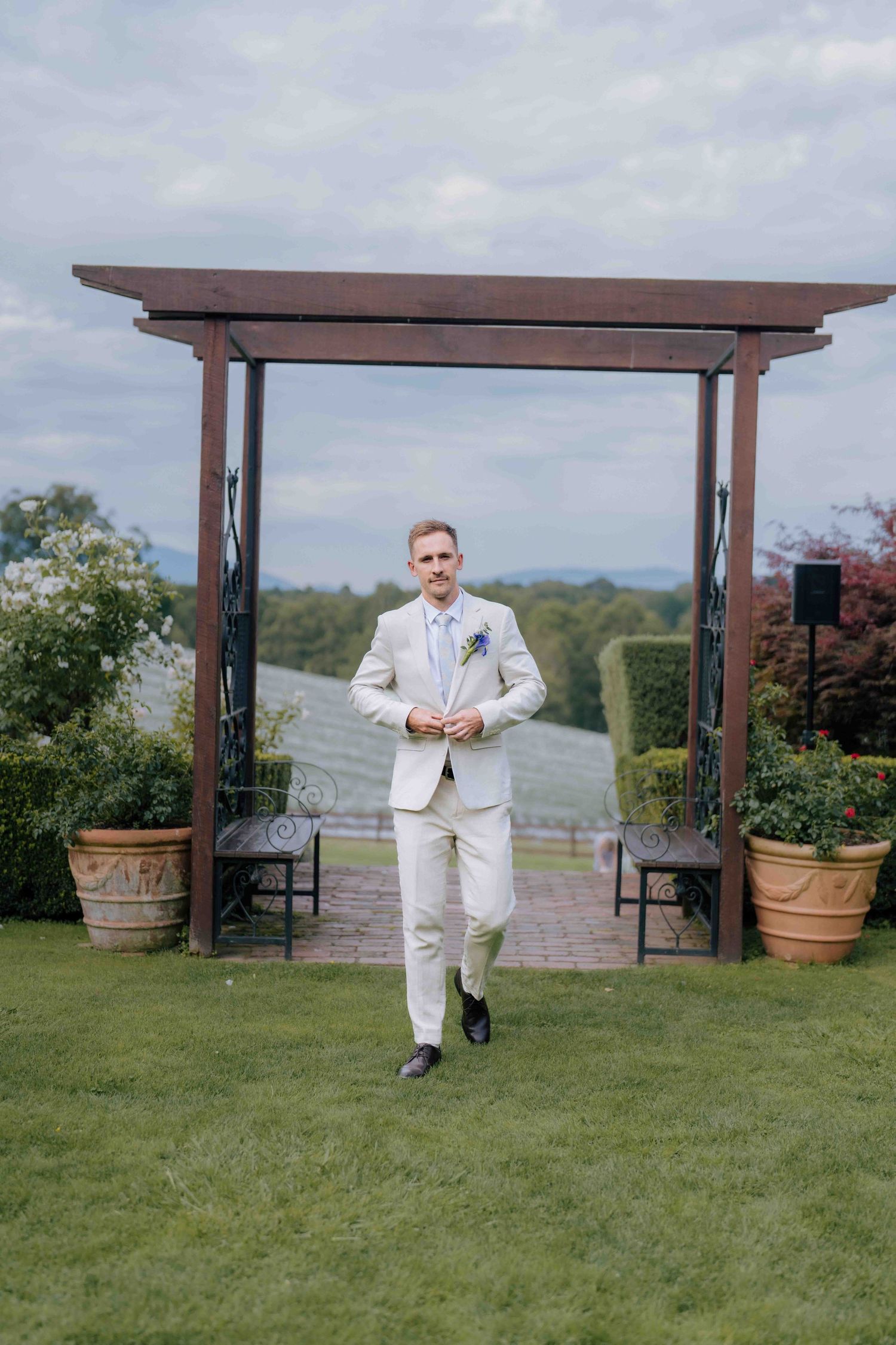 Groom in cream suit walks across green lawn toward wooden pergola at wedding venue.
