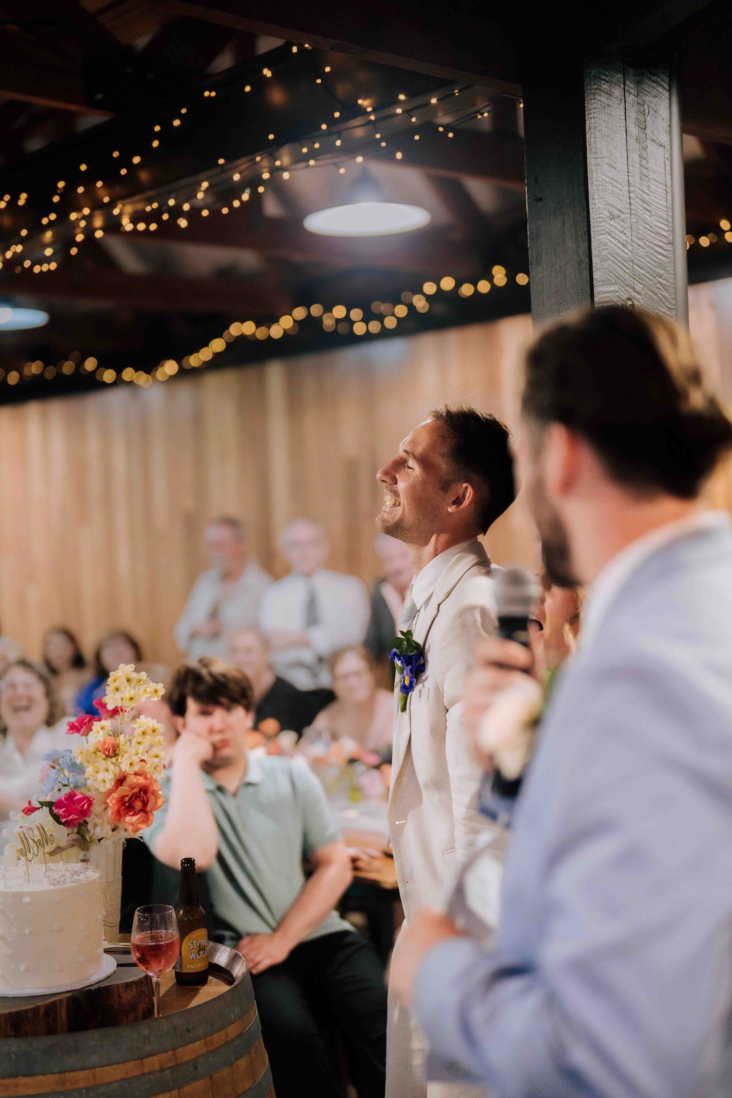 Groom smiles during wedding reception speeches beside a decorated cake on a barrel.