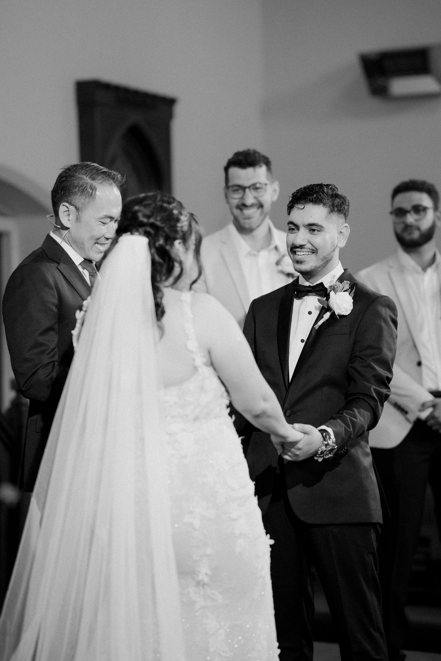 Black and white photo of groom smiling while holding bride's hands during wedding ceremony.