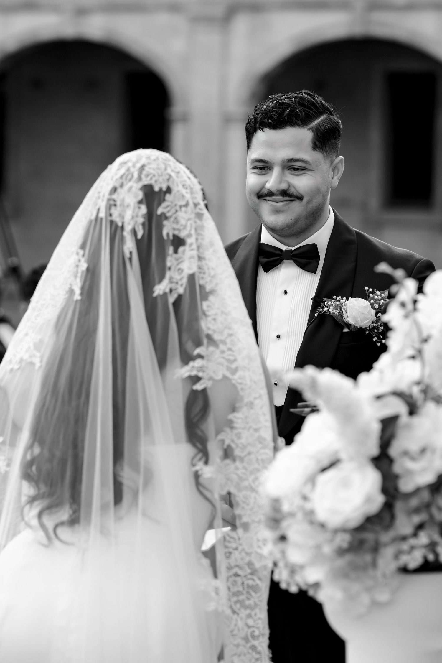 Black and white photo of groom smiling adoringly at bride wearing an ornate lace veil.