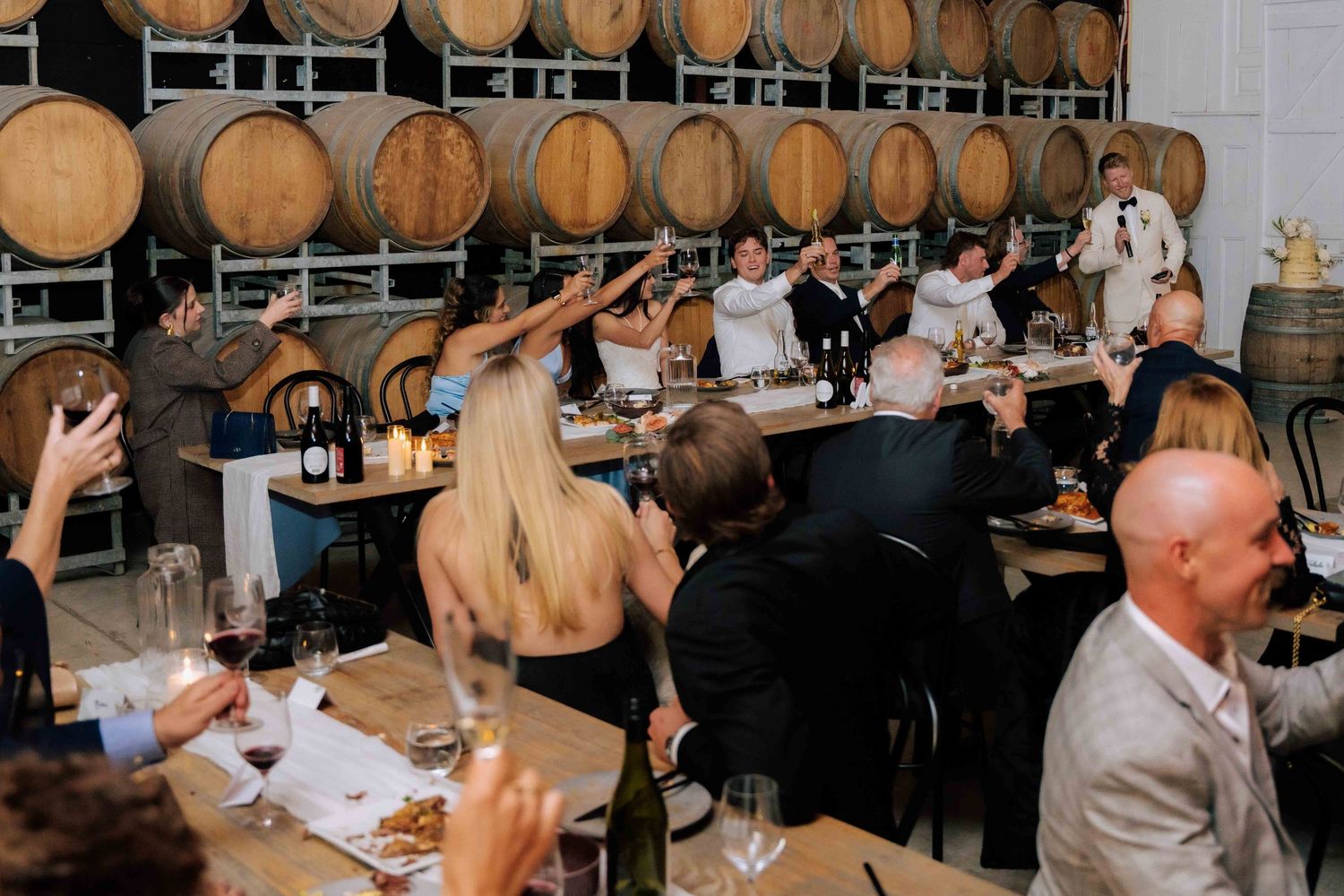 Guests raising glasses in a toast at a winery wedding reception with oak barrels lining the walls.